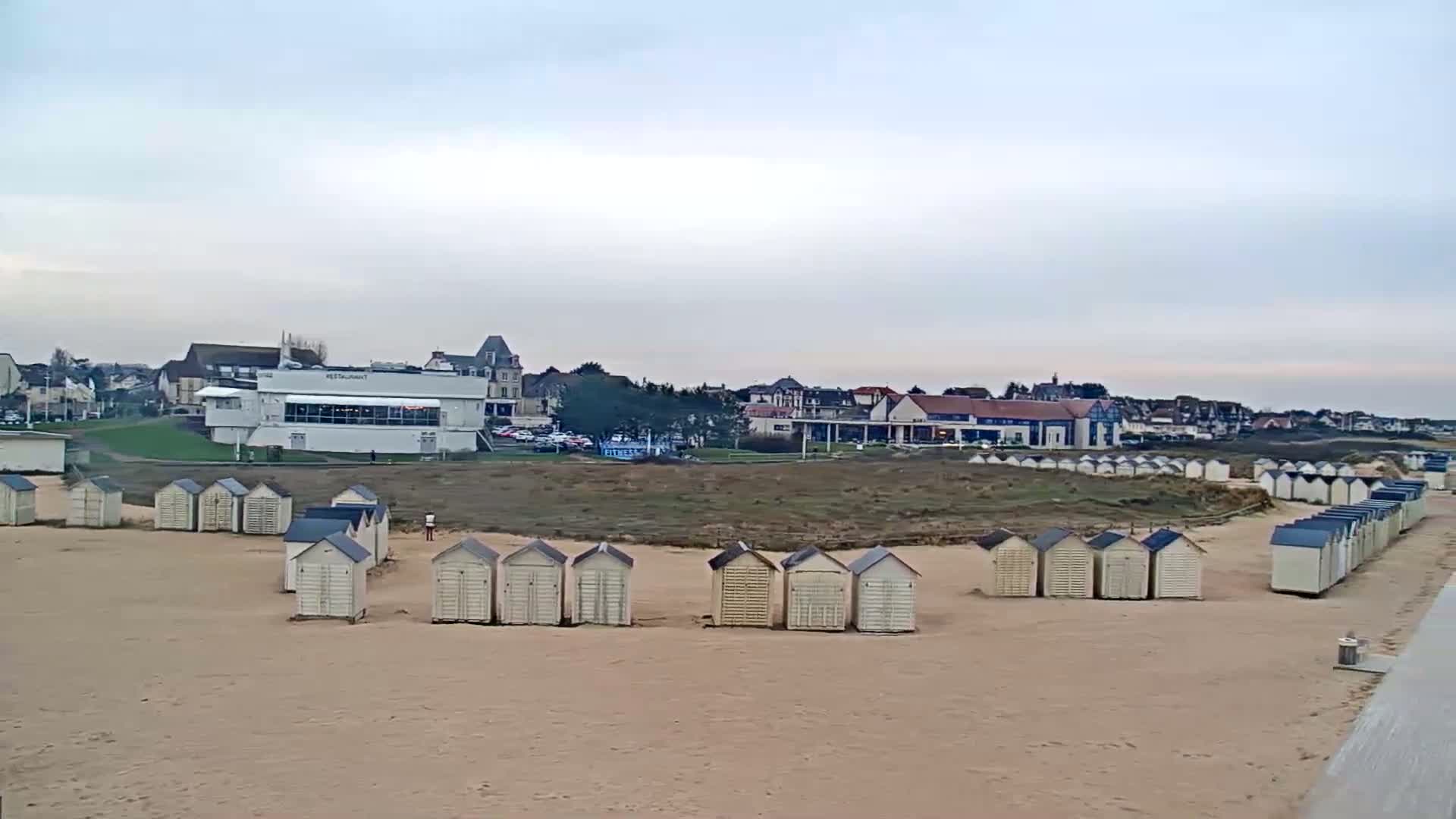 Rows of beach huts sit on a sandy beach in front of a line of buildings under a cloudy sky.
