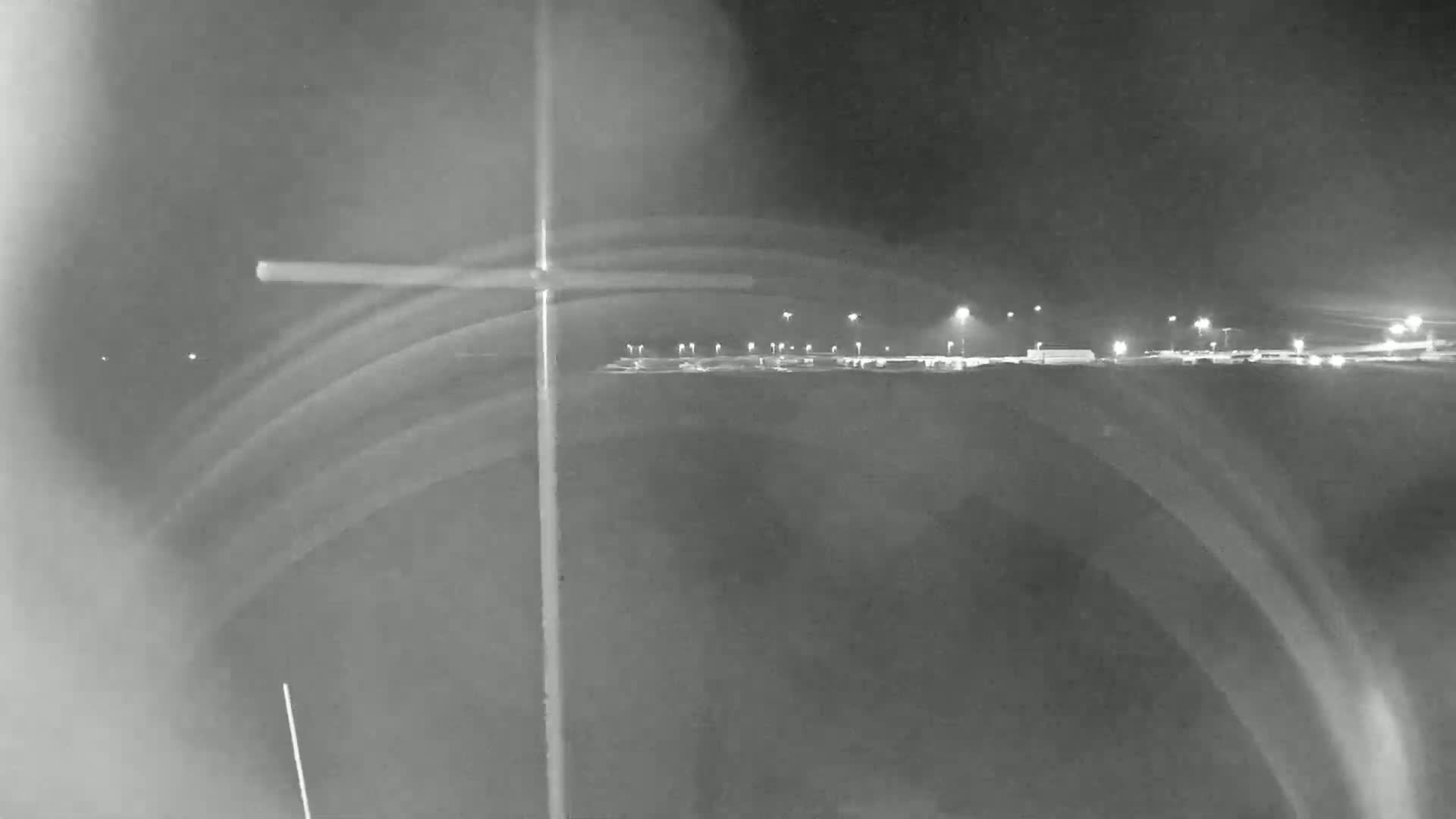A nighttime, low-light image shows a dimly lit parking lot in the distance with several lights, and a tall pole in the foreground with blurry streaks that appear to be caused by motion or light trails.