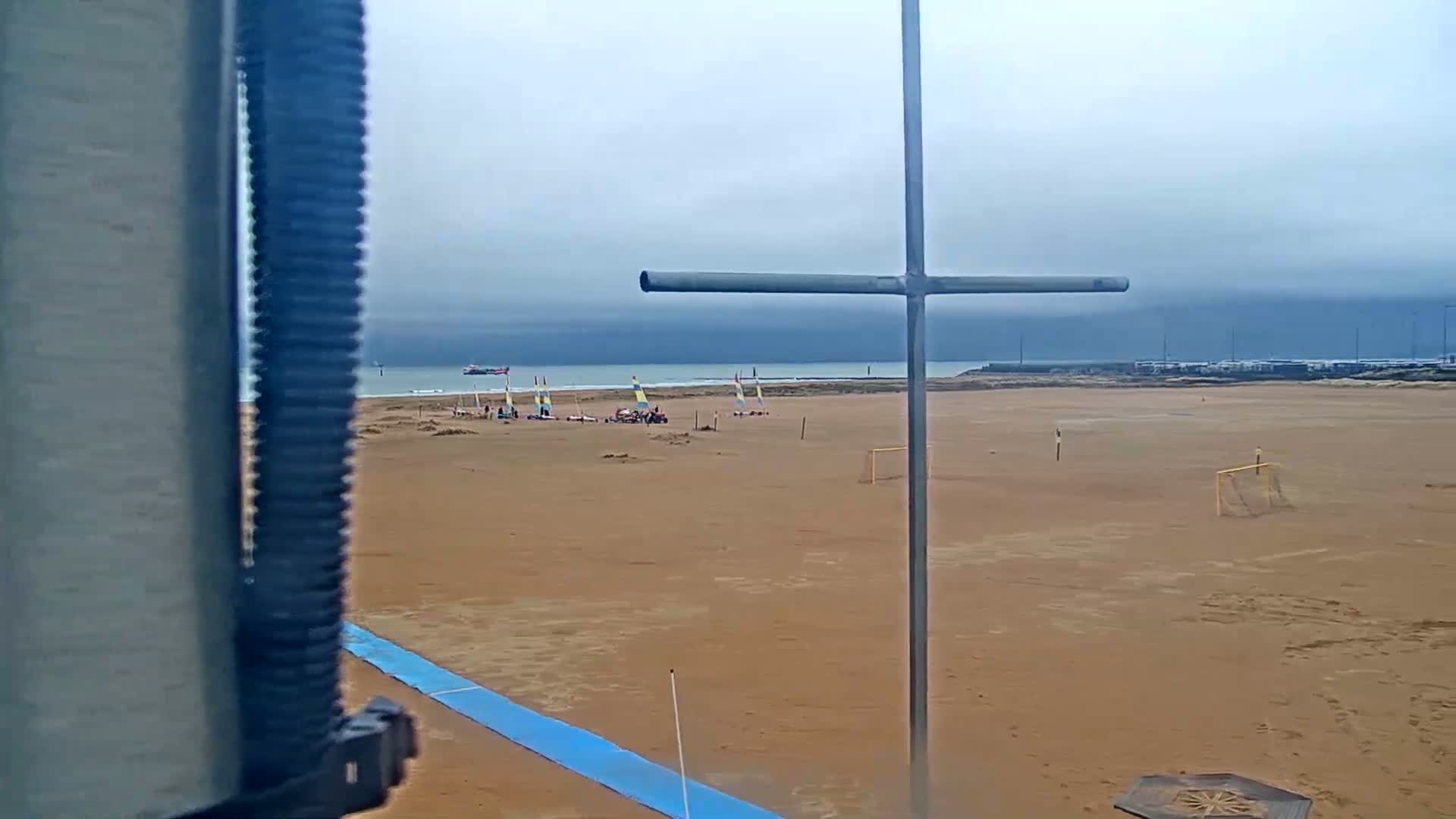 A sandy beach with several sailboats and a soccer goal is visible under an overcast sky.