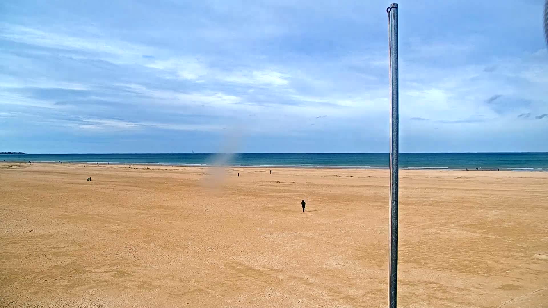 A mostly empty sandy beach stretches to a calm ocean under a partly cloudy sky, with a small dust devil visible on the sand and a lone person walking in the distance.