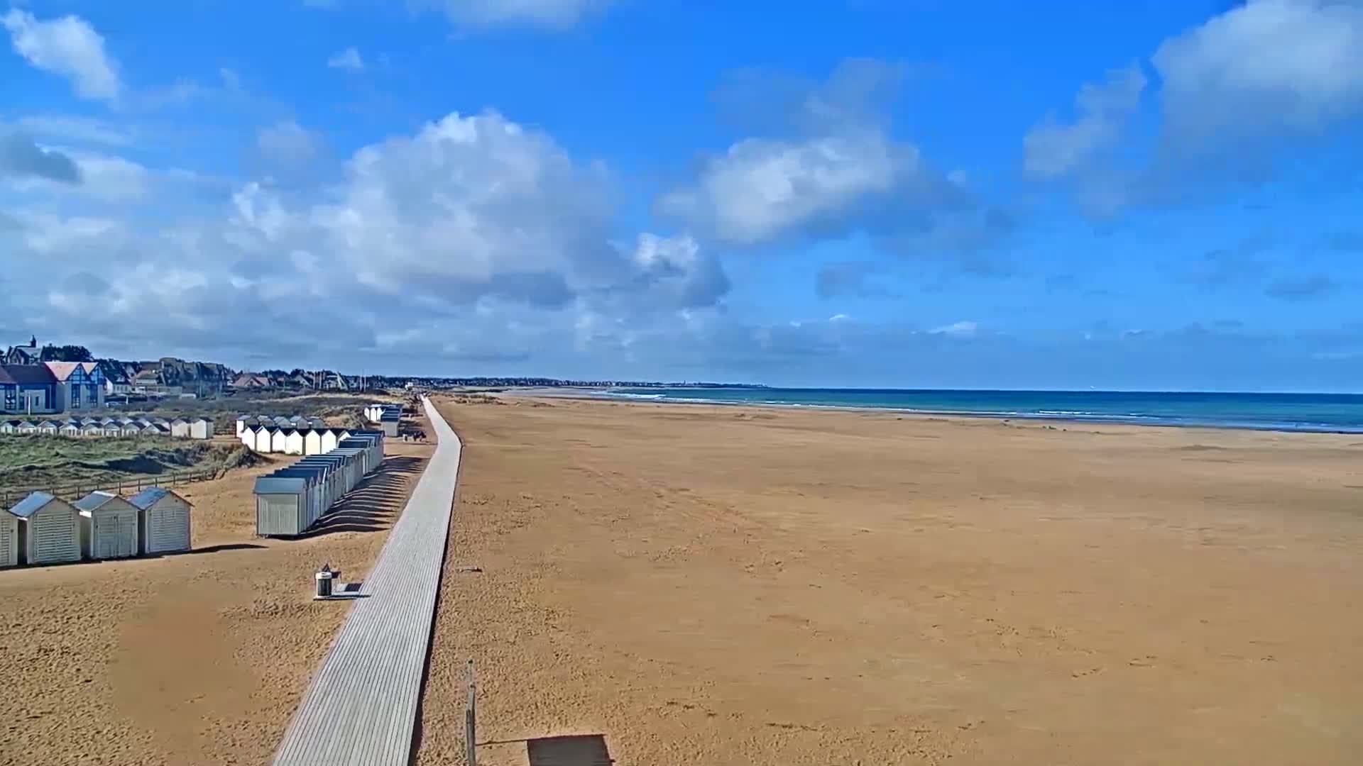 A wide sandy beach stretches to a calm ocean under a partly cloudy blue sky, with rows of beach huts and a boardwalk leading from a town in the background.