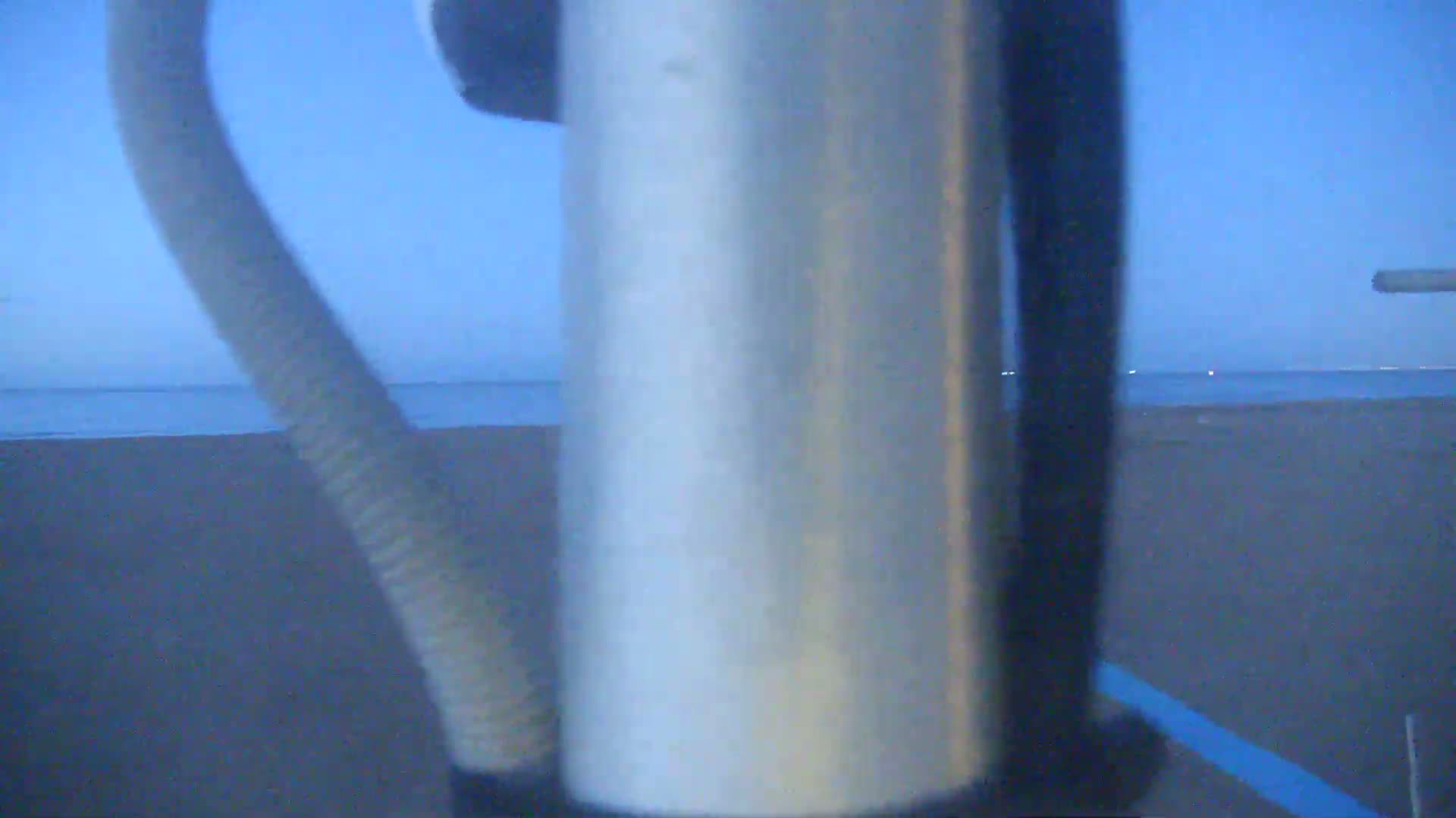 A metal pole is in the foreground, partially obscuring a view of a calm ocean and dark sandy beach under a twilight sky.