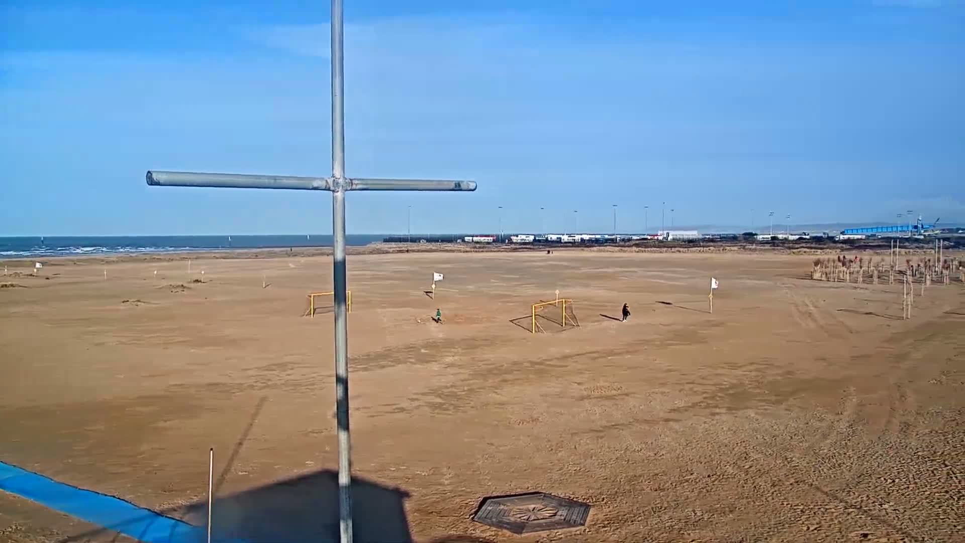 A sandy beach soccer field with two goals and a few people playing under a clear blue sky.