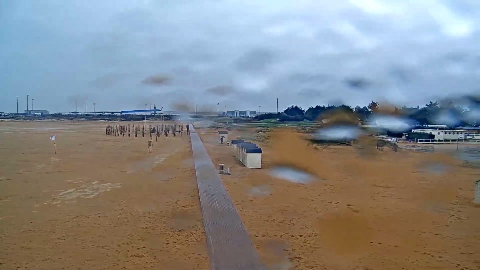 A long boardwalk stretches across a wet, sandy beach toward a distant airport under an overcast sky.