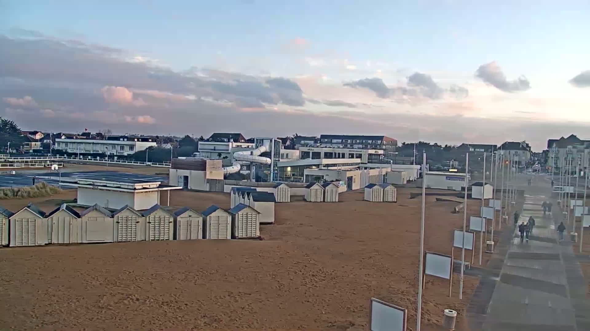 A sandy beach with rows of beach huts, a building with a waterslide, and a paved walkway with people walking under a mostly cloudy sky.