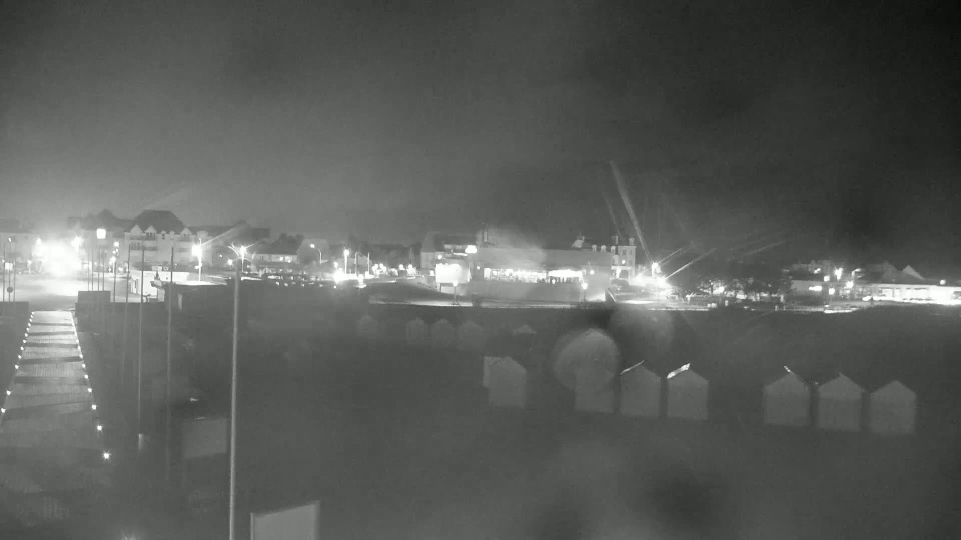 A nighttime, rain-streaked view of a beach town with illuminated buildings and rows of beach huts in the foreground.