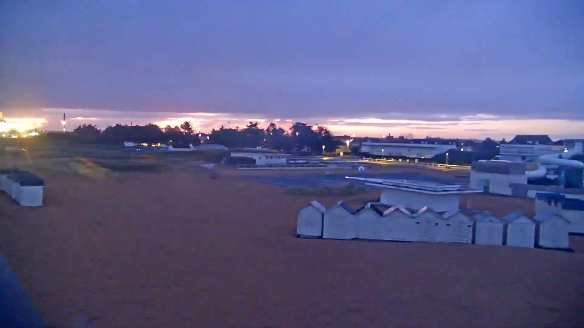 Under a twilight sky, a sandy beach features rows of small white beach huts near buildings and a swimming pool.