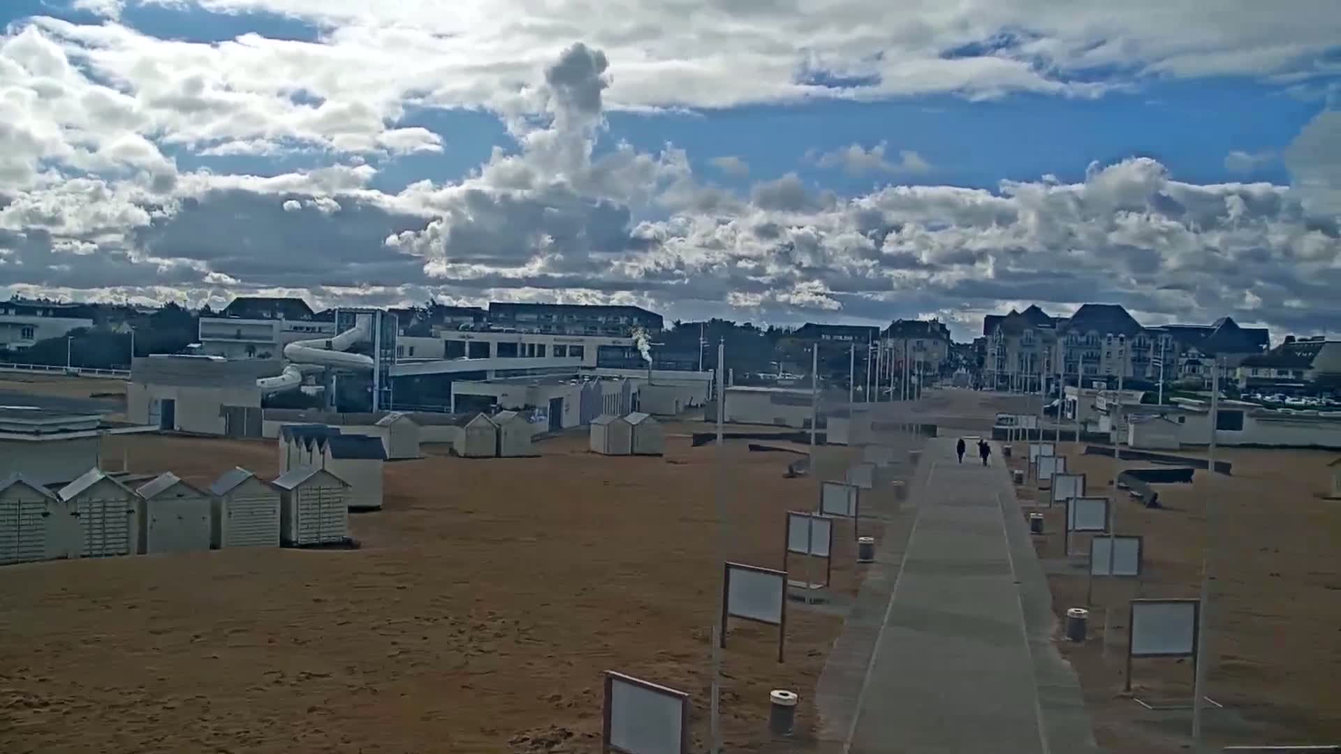 A sandy beach with beach huts, a water slide, and buildings in the background under a partly cloudy sky.
