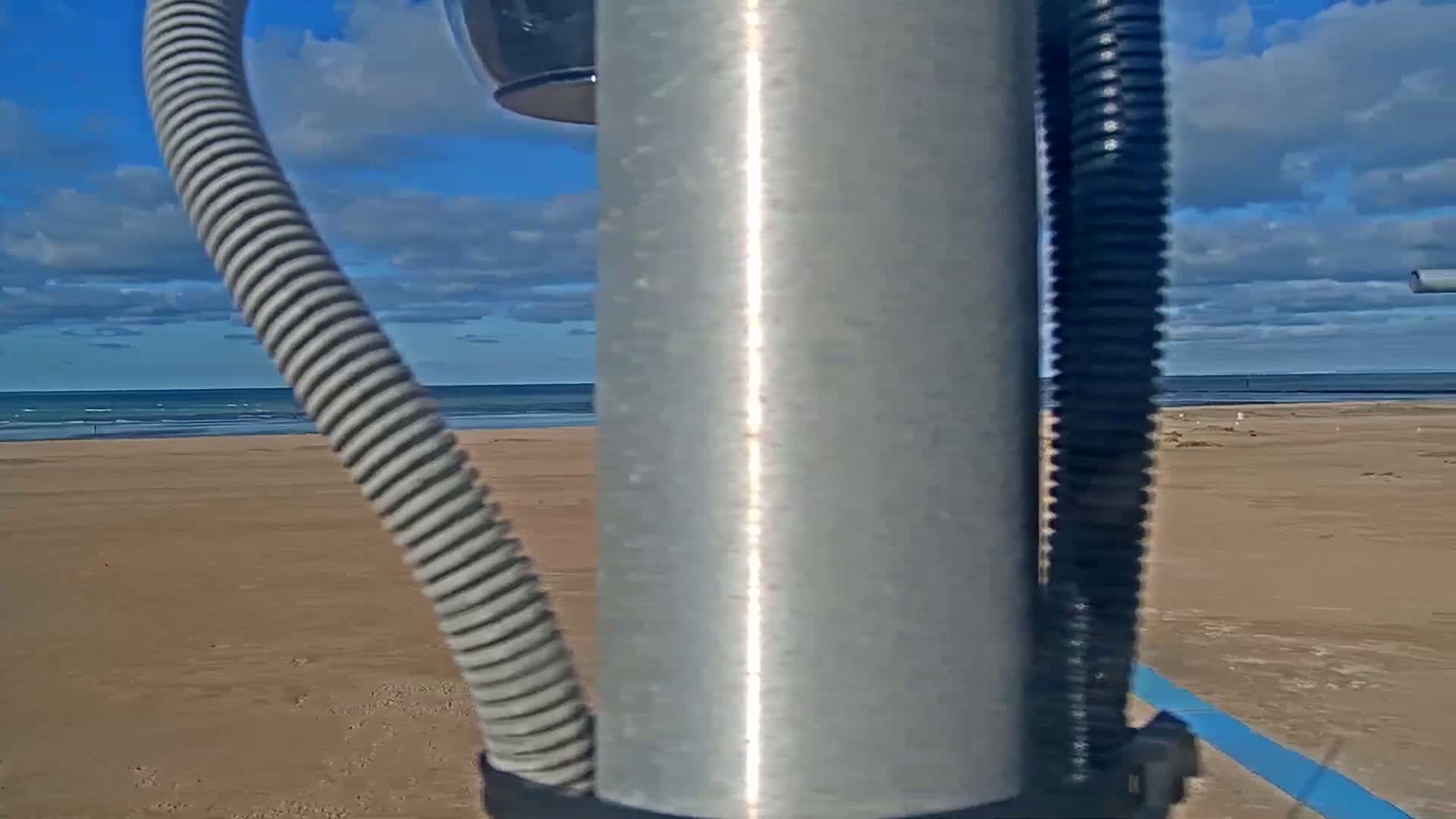 A mostly sunny day shows a sandy beach and ocean viewed from behind a metal pole with attached cords and a camera.