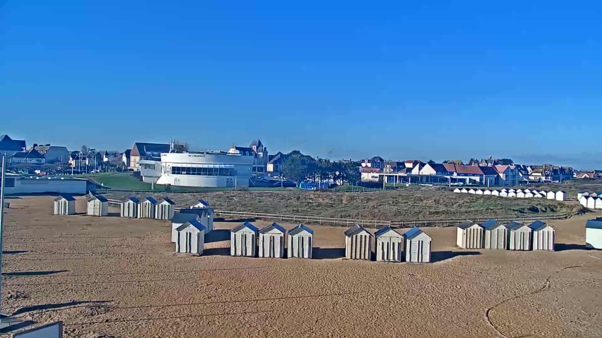 A sandy beach with rows of white beach huts and a large white building behind it, under a clear blue sky.