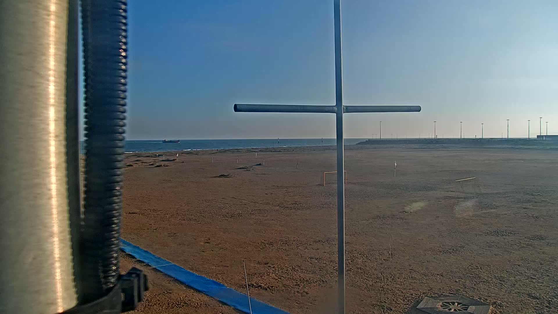 A sandy beach with a metal cross-shaped structure in the foreground, under a clear blue sky, and a calm ocean in the background.