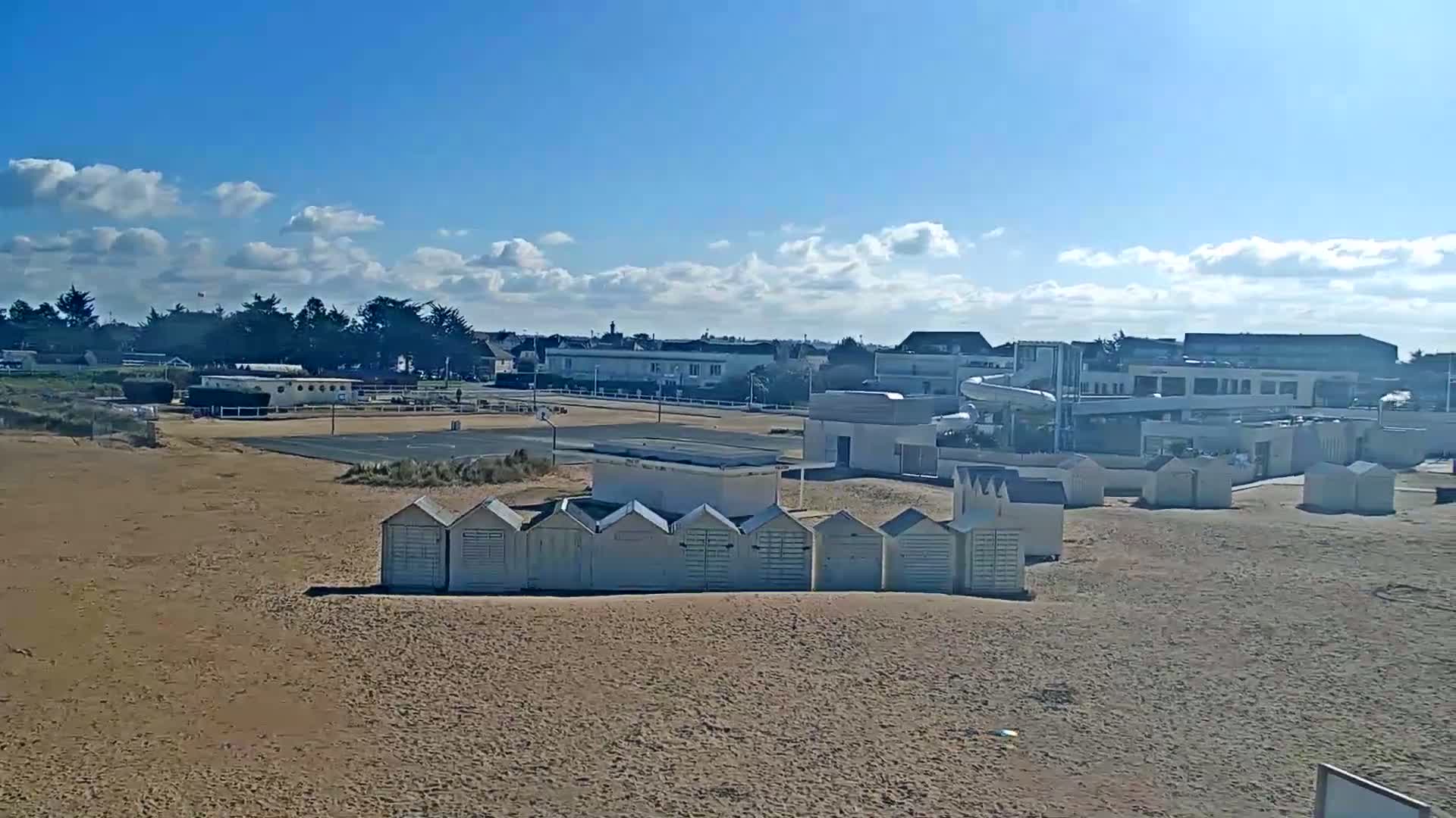 A sandy beach with rows of beach huts, a water slide, and buildings in the background under a partly cloudy blue sky.