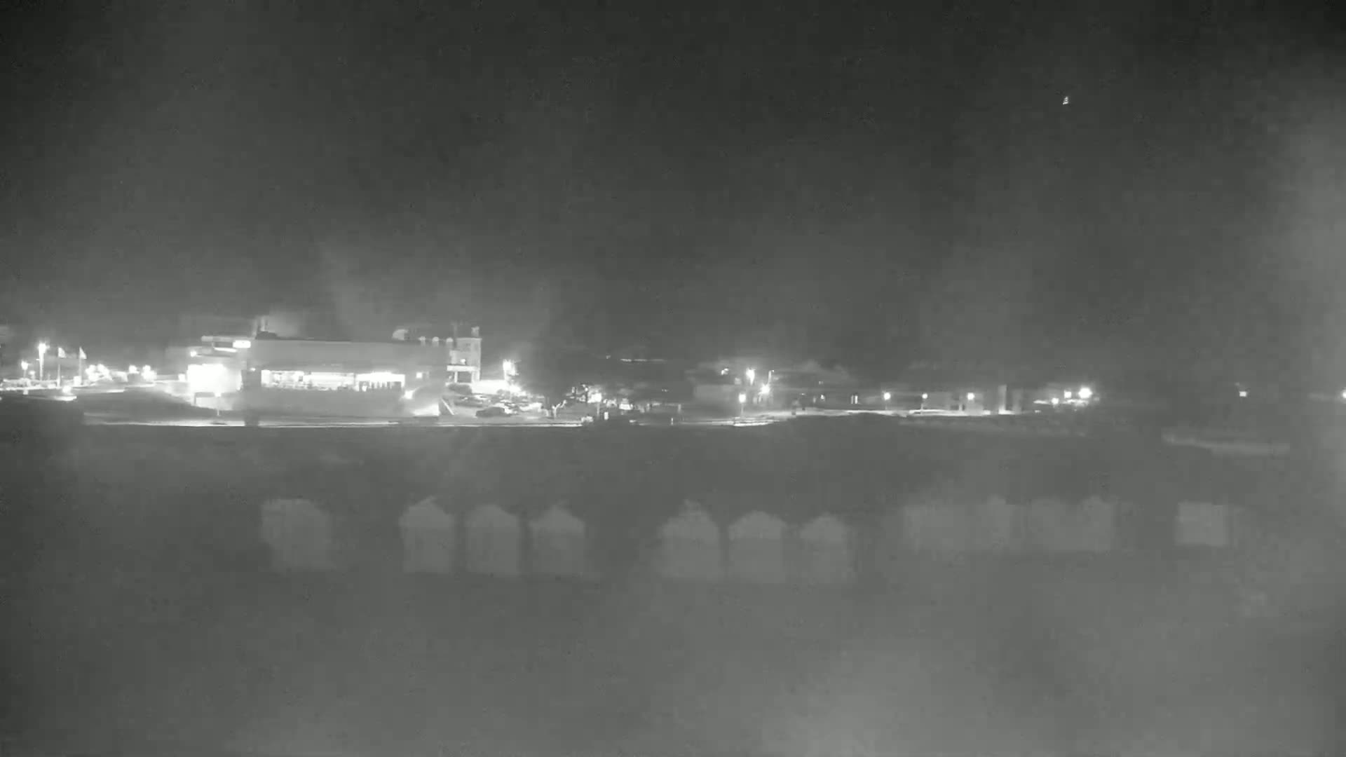 A nighttime, low-light view shows a row of beach huts in the foreground, with a building and some lights visible in the background under a dark, clear sky.