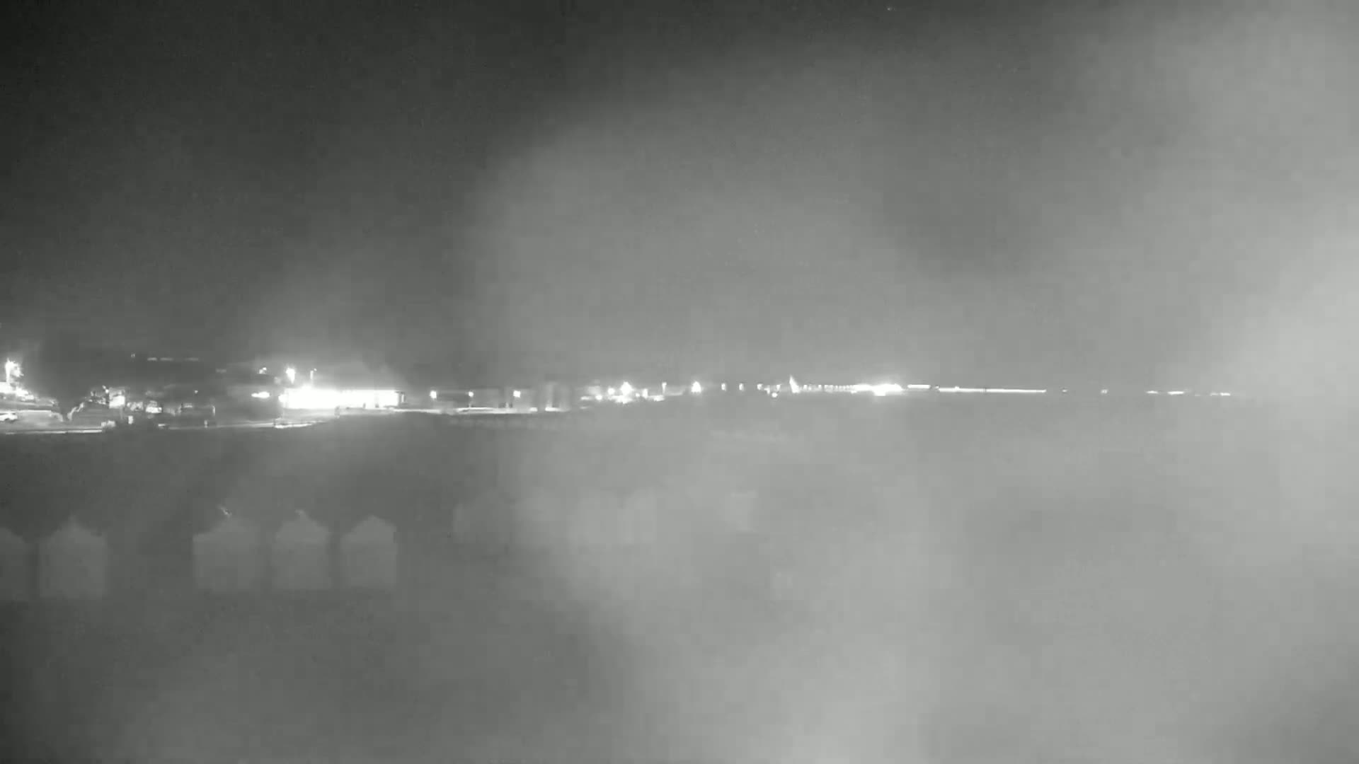 A nighttime view shows a foggy or hazy scene with distant lights of a town or city visible over what looks like a low wall or barrier.