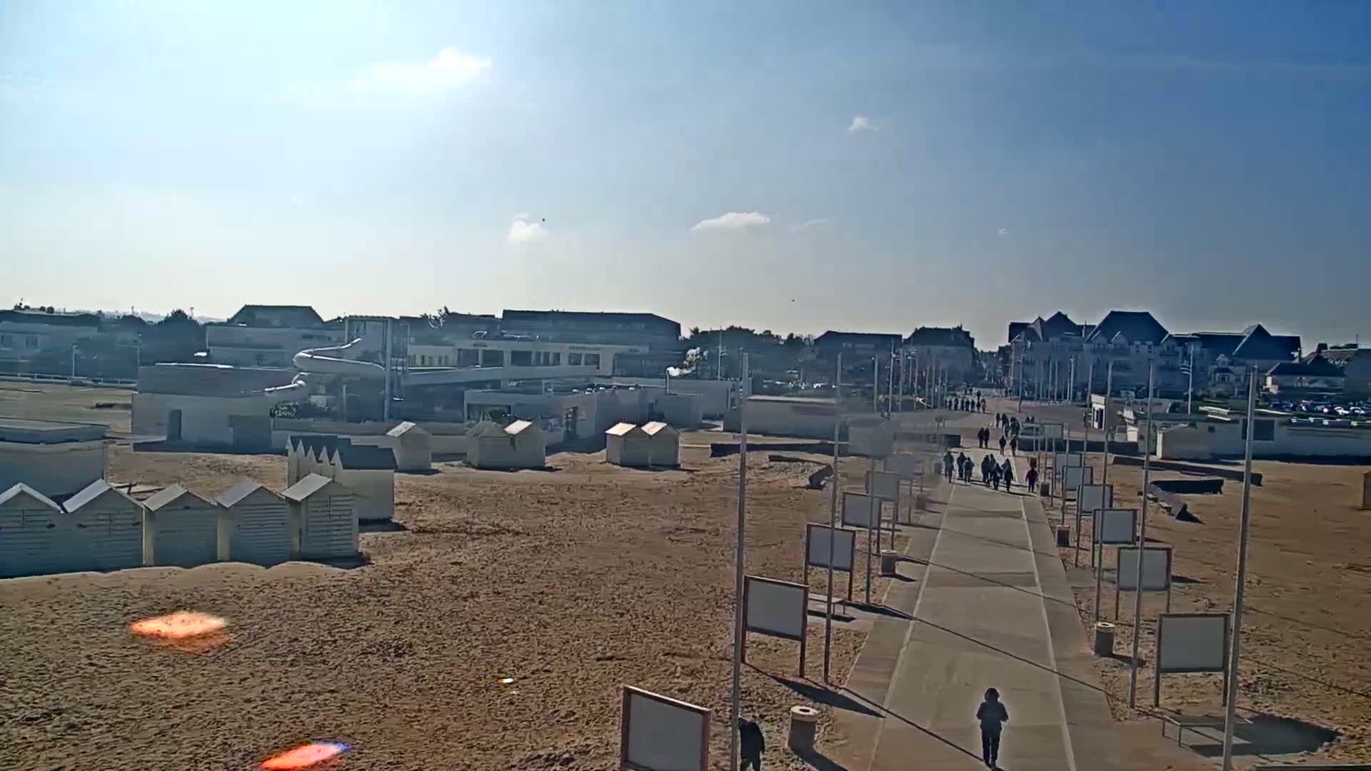A sandy beach with rows of small beach huts, a paved walkway with people strolling, and a water park structure in the background under a sunny sky.