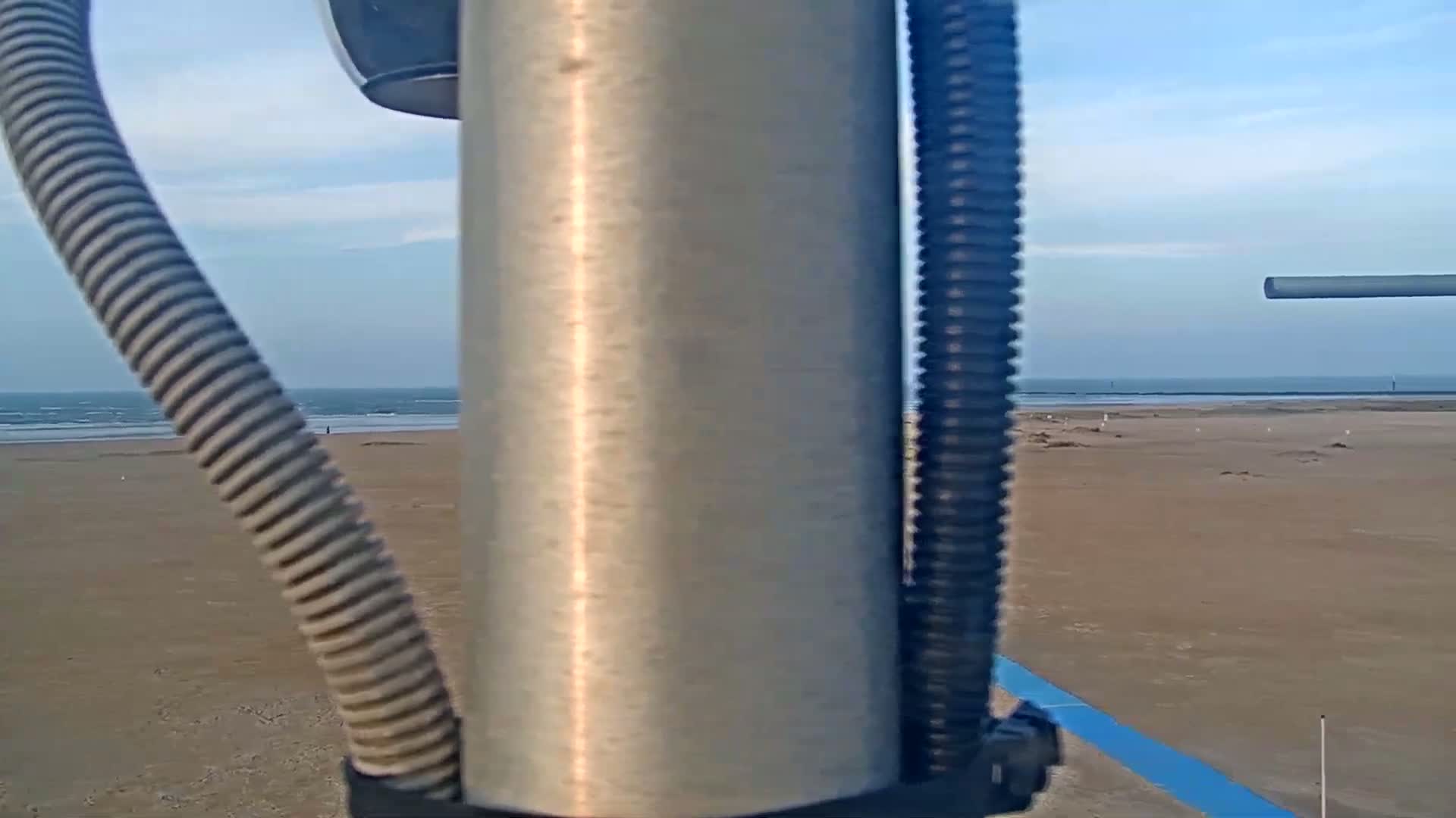 A metal pole with cords attached is in the foreground, partially obscuring a view of a sandy beach and calm ocean under a mostly clear sky.