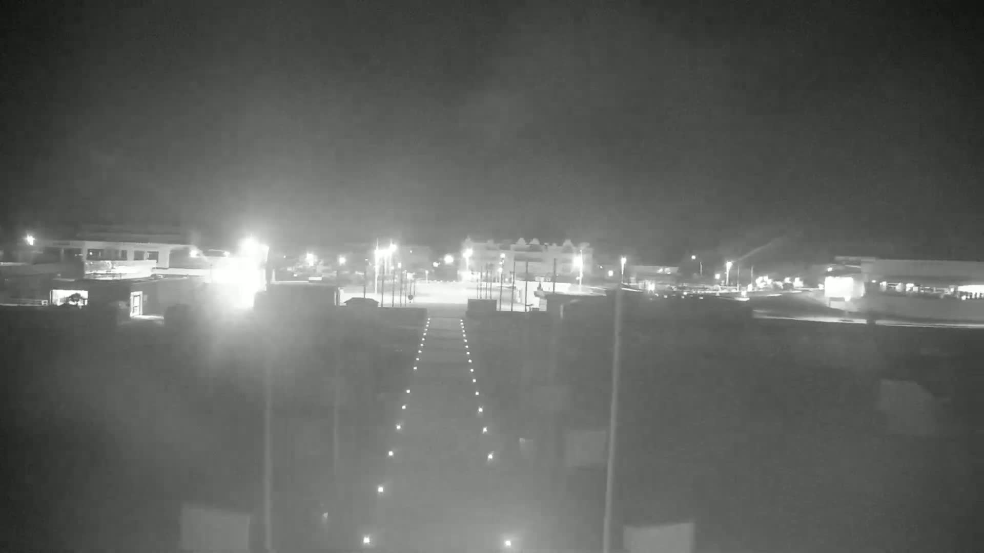 A nighttime view of a paved area with buildings and streetlights in hazy or foggy conditions.