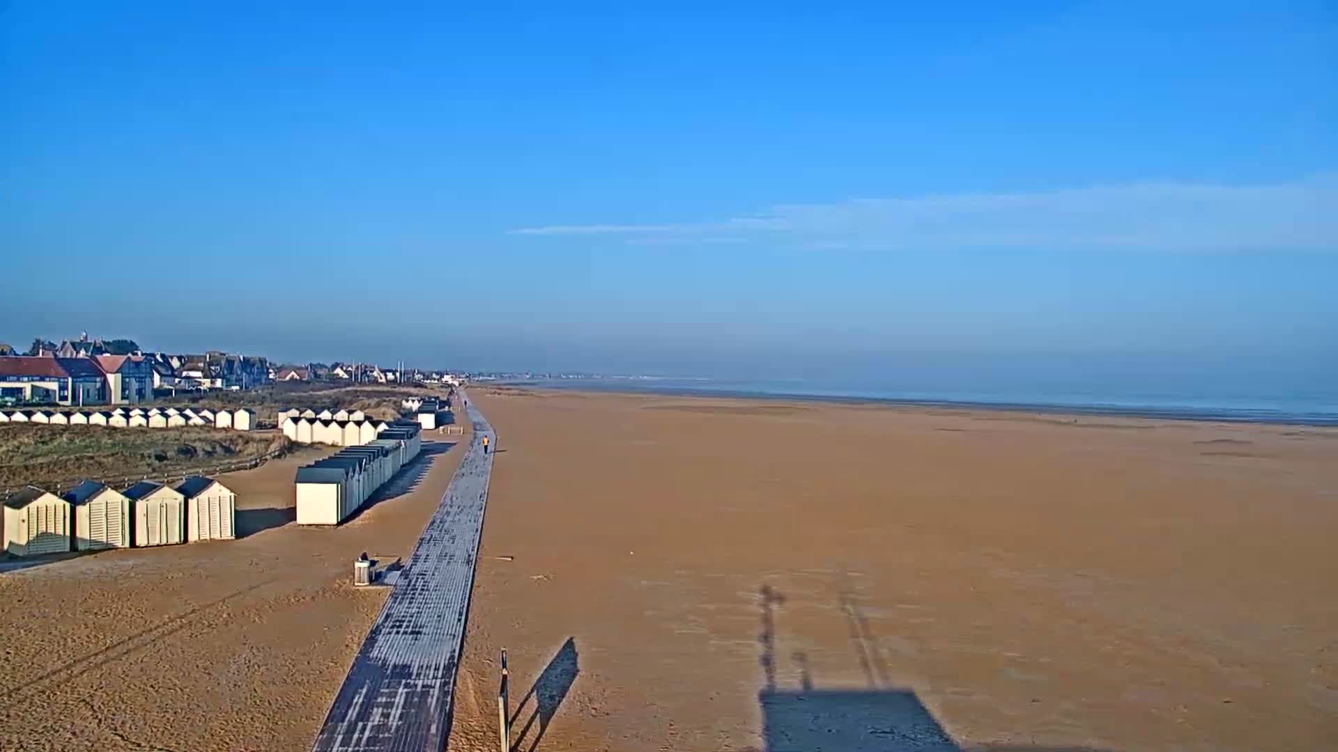 A sandy beach stretches to a calm ocean under a clear blue sky, with beach huts and a boardwalk lining the shore near buildings in the distance.