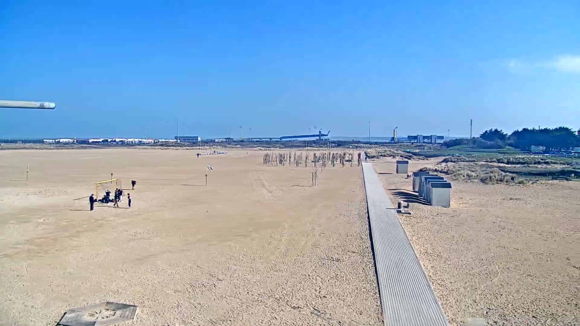 A sandy beach on a sunny day features a group of people playing soccer, a boardwalk, beach huts, and a distant view of a harbor.