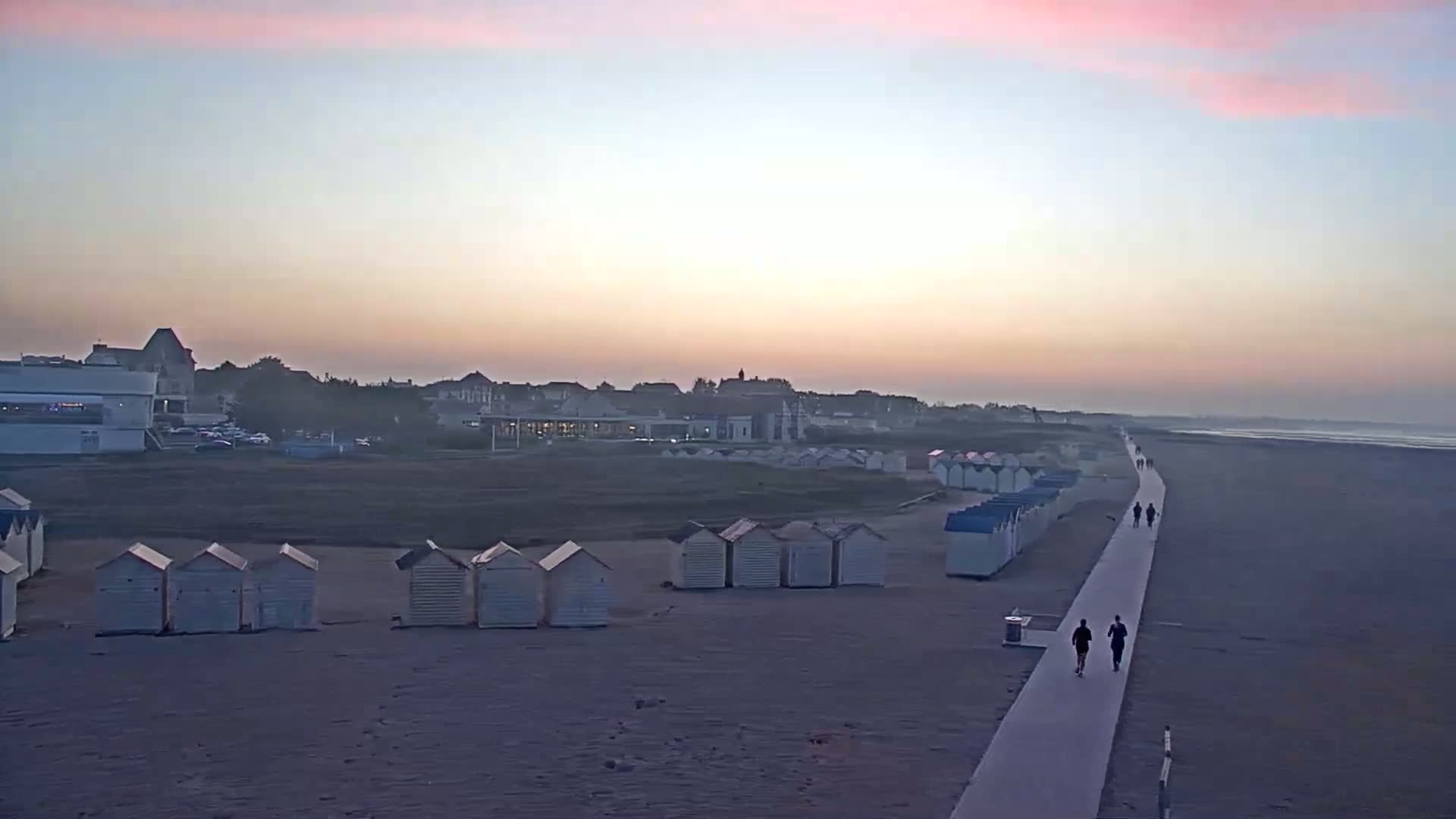 A sandy beach at sunrise, with rows of small beach huts and people walking along a boardwalk towards the ocean.