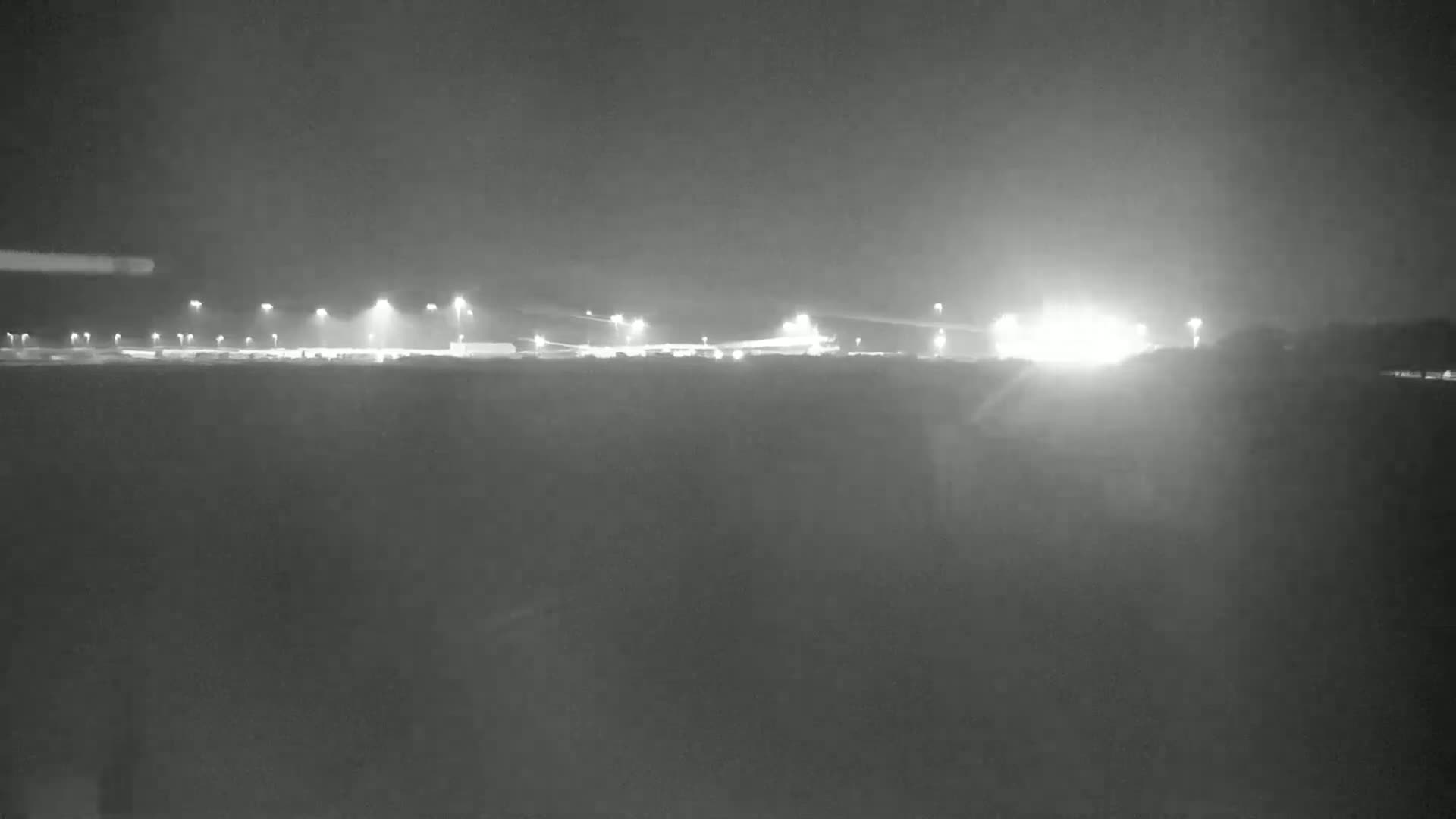 A nighttime, low-light view of a brightly lit industrial area or airport across a dark, flat expanse of land.