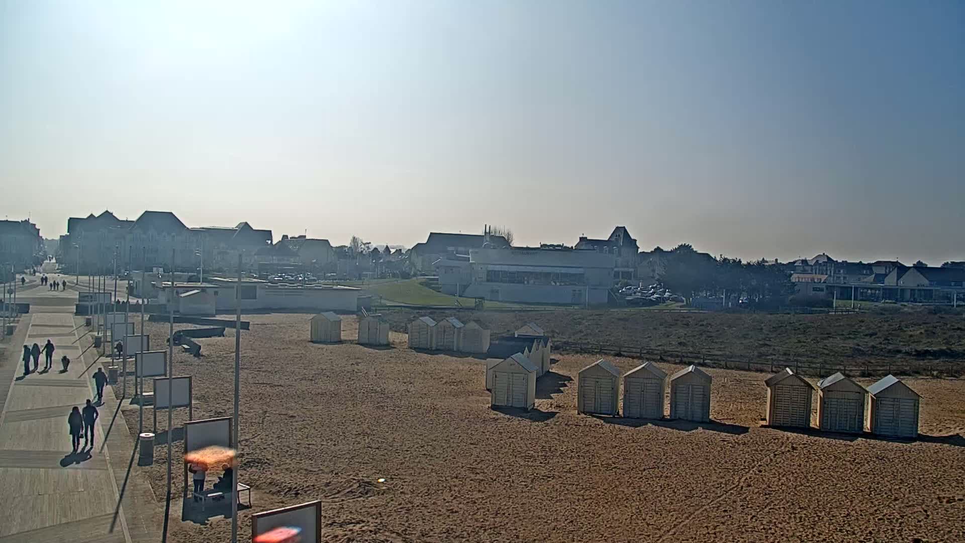 A sandy beach with rows of beach huts is visible in front of a town under a clear, sunny sky.