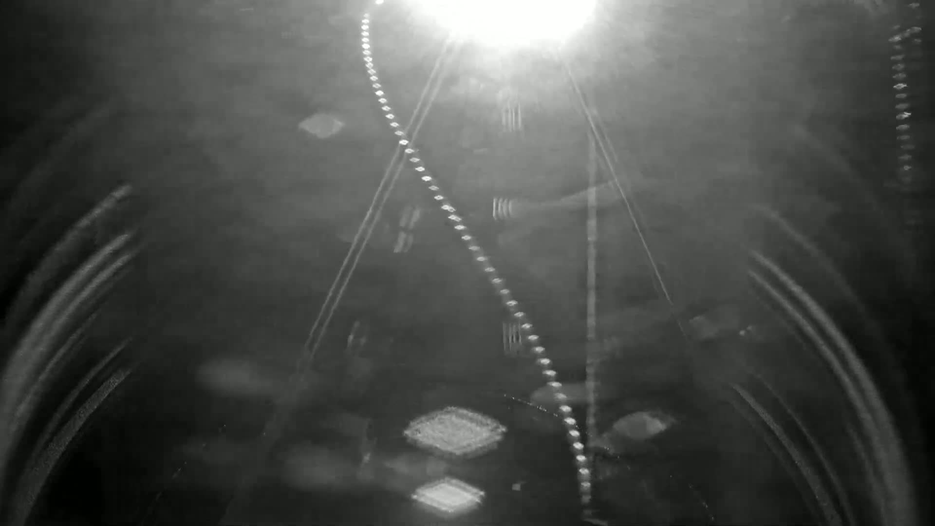 A blurry, nighttime image shows a bright light in the distance with streaks of light suggesting movement, possibly from a vehicle's headlights, along a dark road or tunnel.
