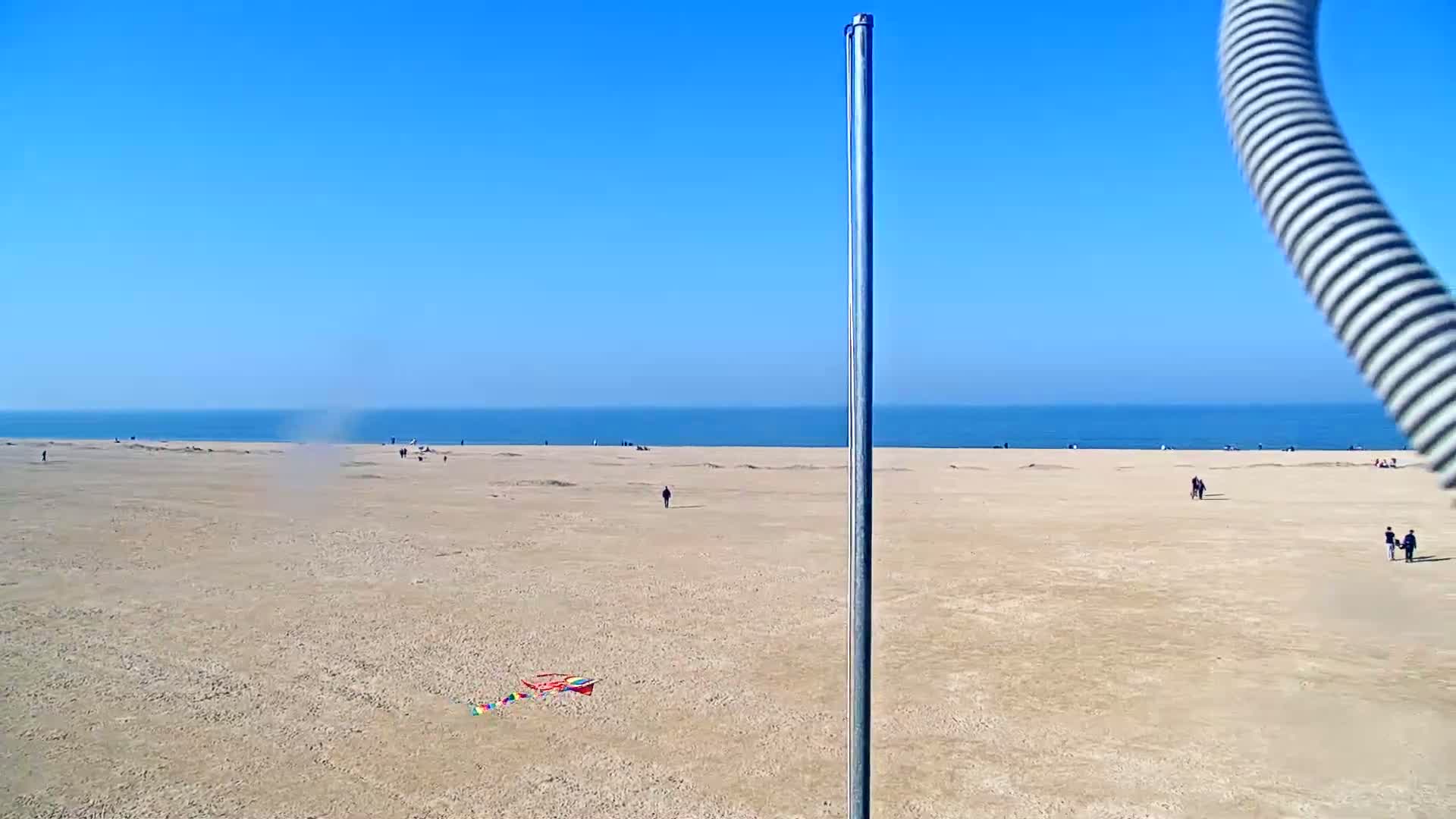 A wide sandy beach stretches to a calm blue ocean under a clear blue sky, with a few people scattered across the beach and a small kite lying on the sand.