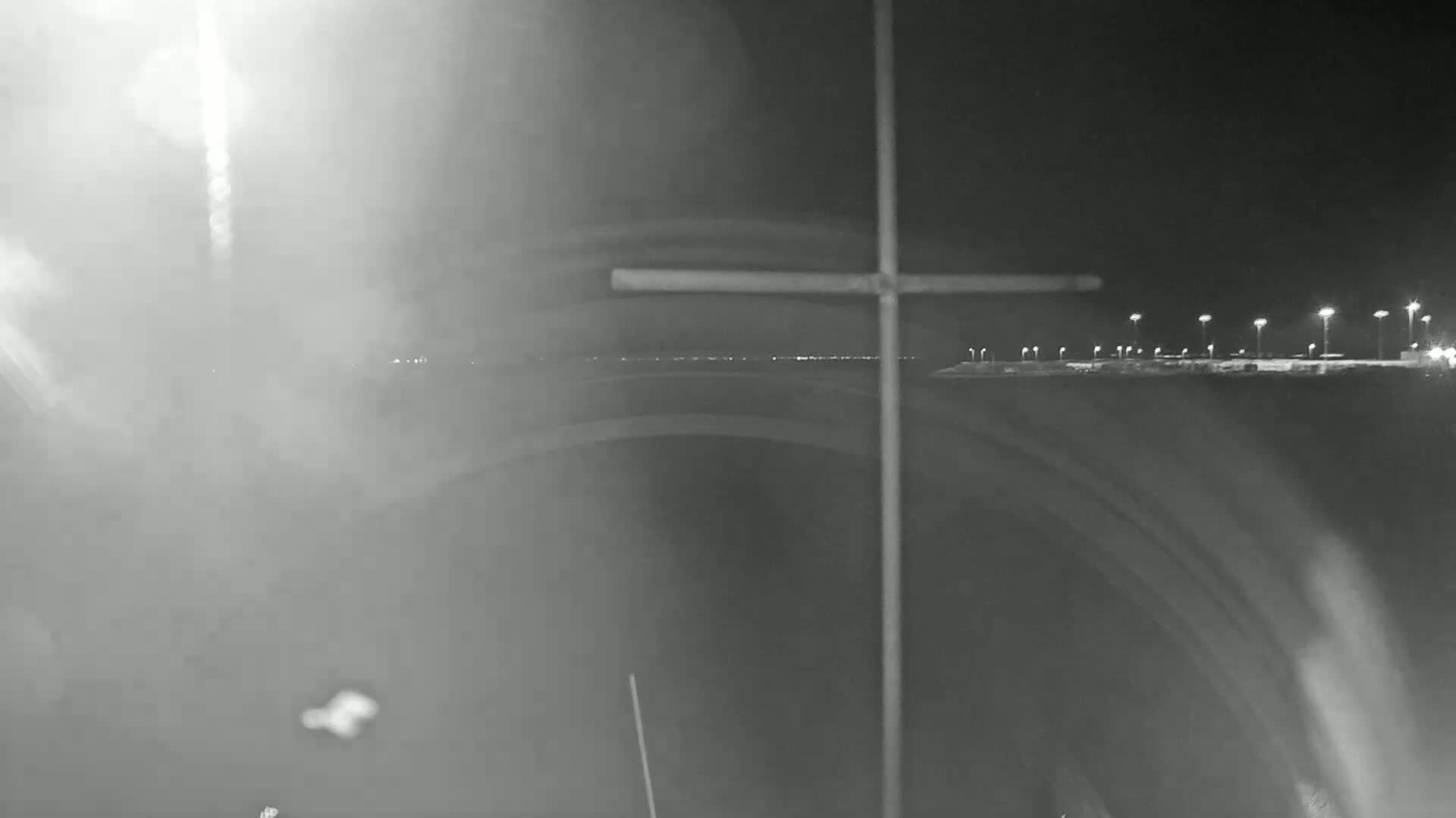 A hazy night scene shows a distant line of lights, possibly a parking lot or roadway, viewed through a curved structure with a cross-shaped object in the foreground.