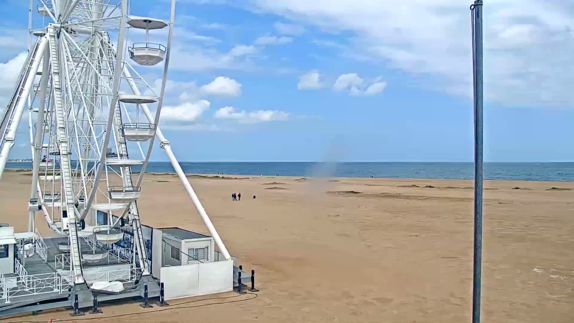 A large white Ferris wheel sits on a sandy beach next to a calm ocean under a partly cloudy sky.