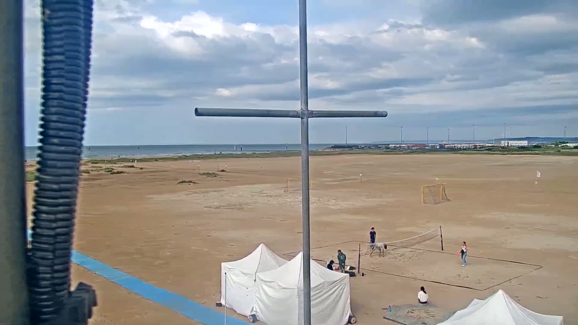 A sandy beach with a volleyball net and soccer goal, two tents, and several people playing sports under a partly cloudy sky.