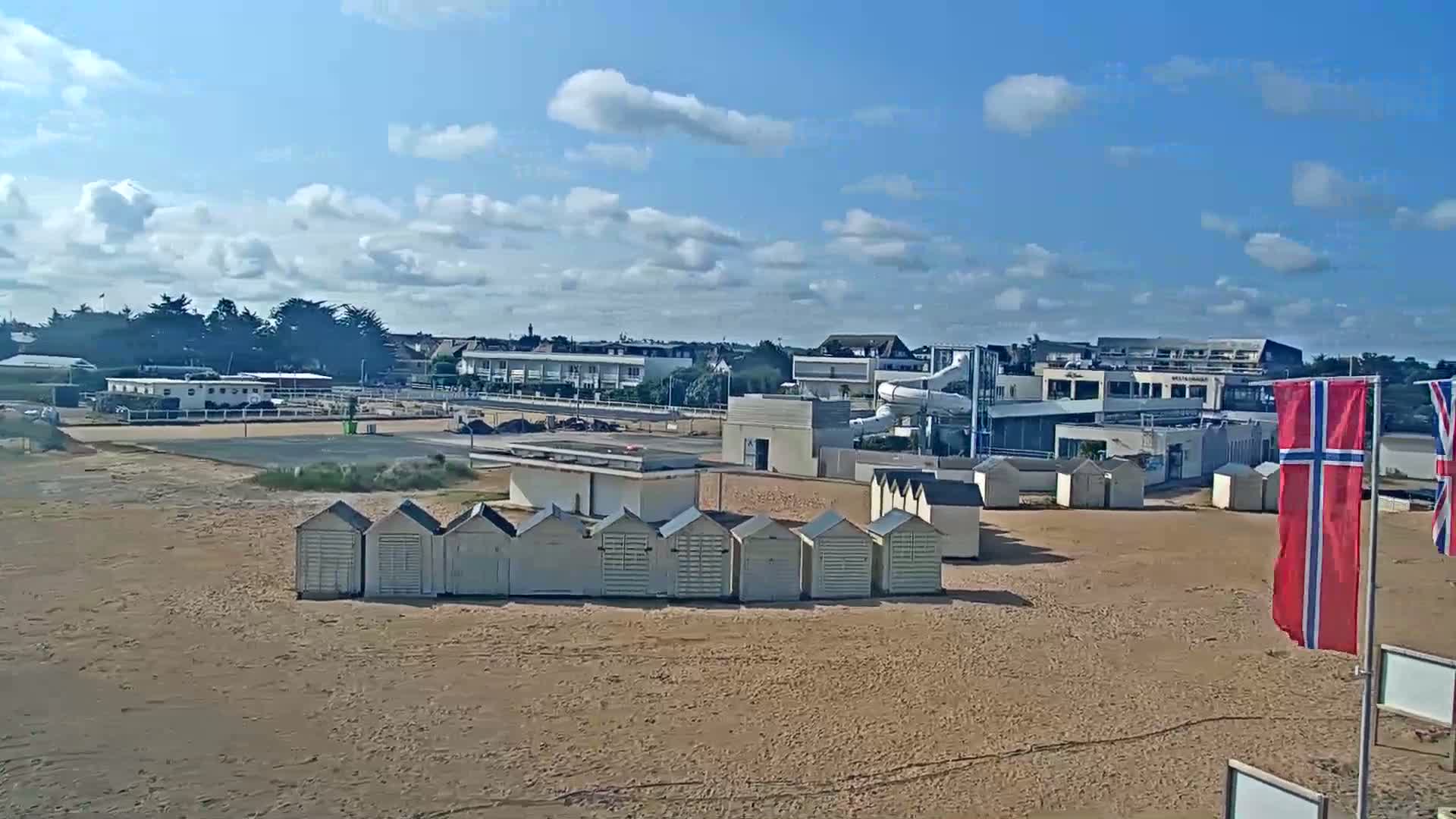 A sandy beach with rows of small beach huts, a water park, and buildings in the background under a partly cloudy sky.