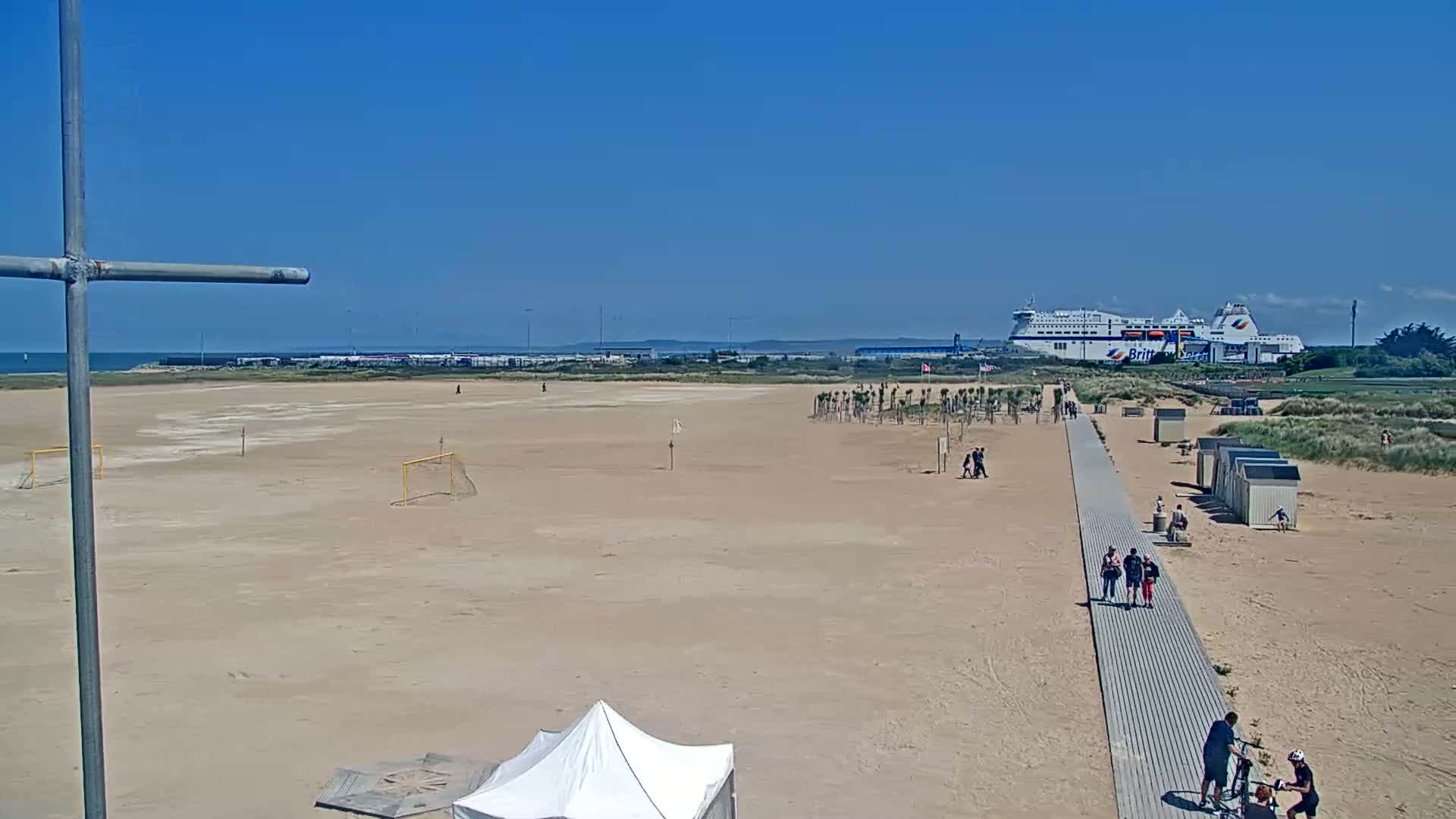 A sandy beach with a boardwalk, beach huts, and soccer goals is shown on a sunny day, with a large ferry visible in the background.