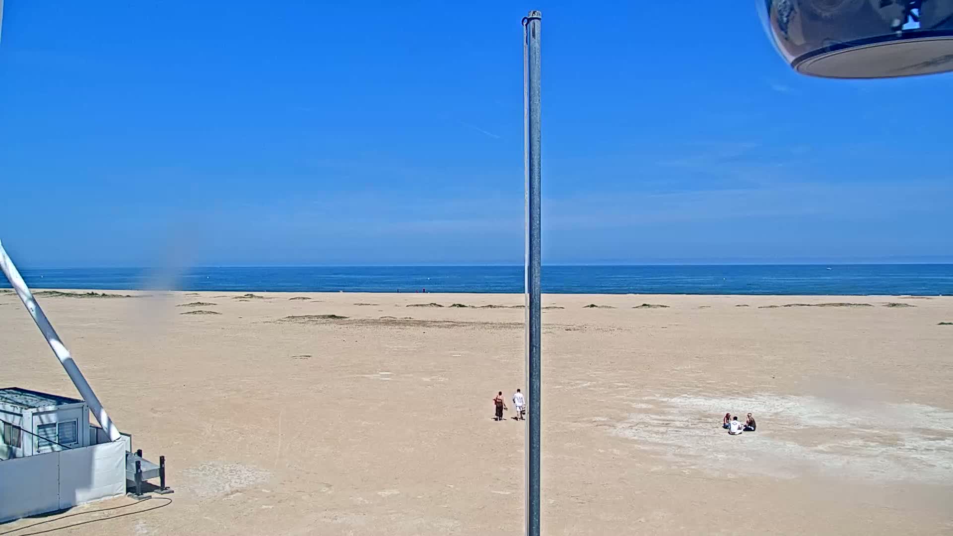 A sandy beach stretches to a calm blue ocean under a clear blue sky, with a few people visible in the distance.
