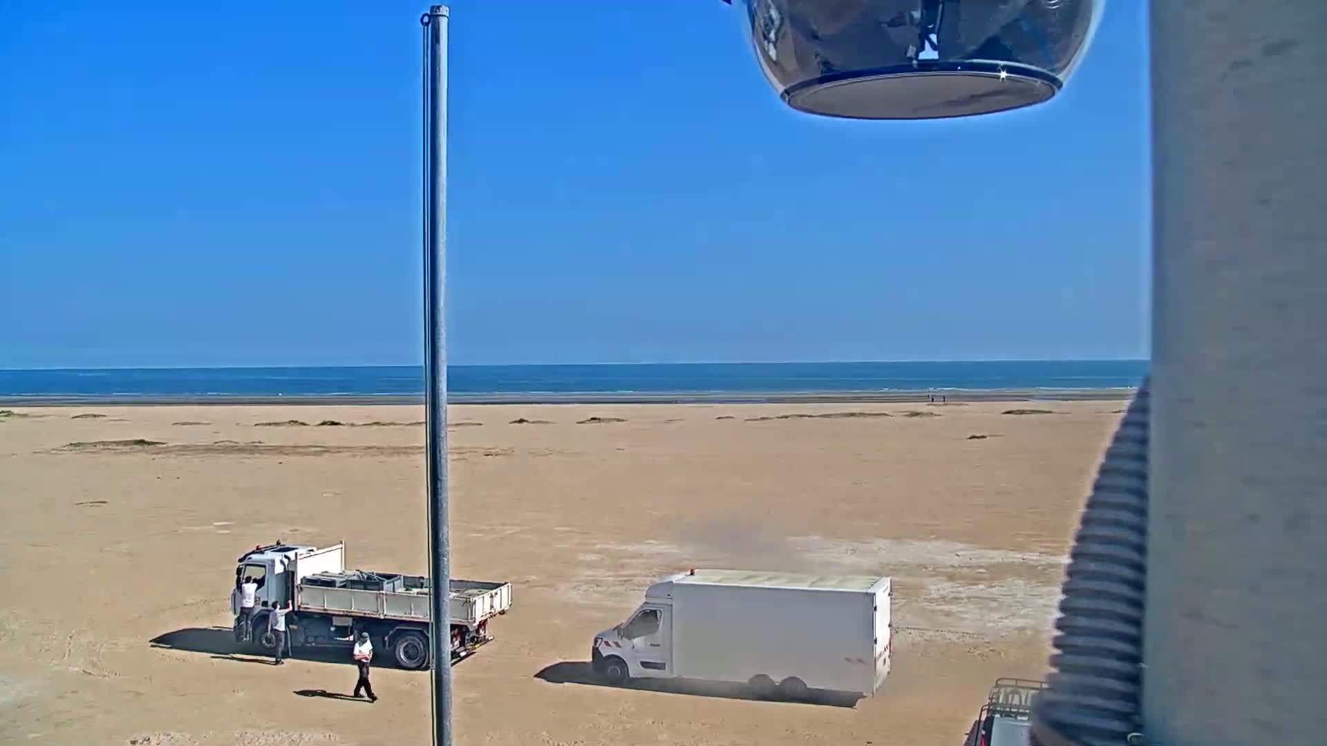 A sandy beach stretches to a calm ocean under a clear blue sky, with a dump truck and a van parked on the sand.