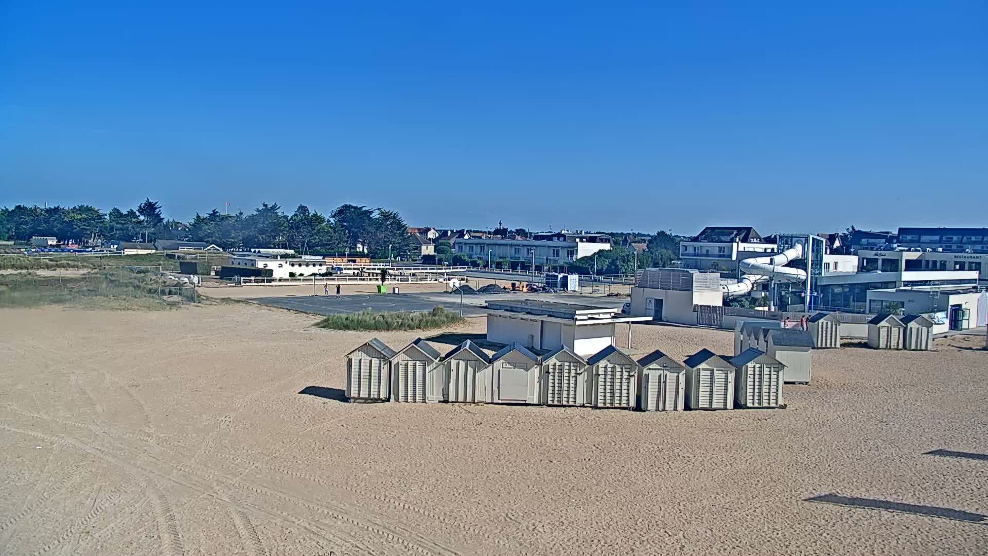 A sandy beach on a clear, sunny day features rows of beach huts, a basketball court, and a water park in the background.