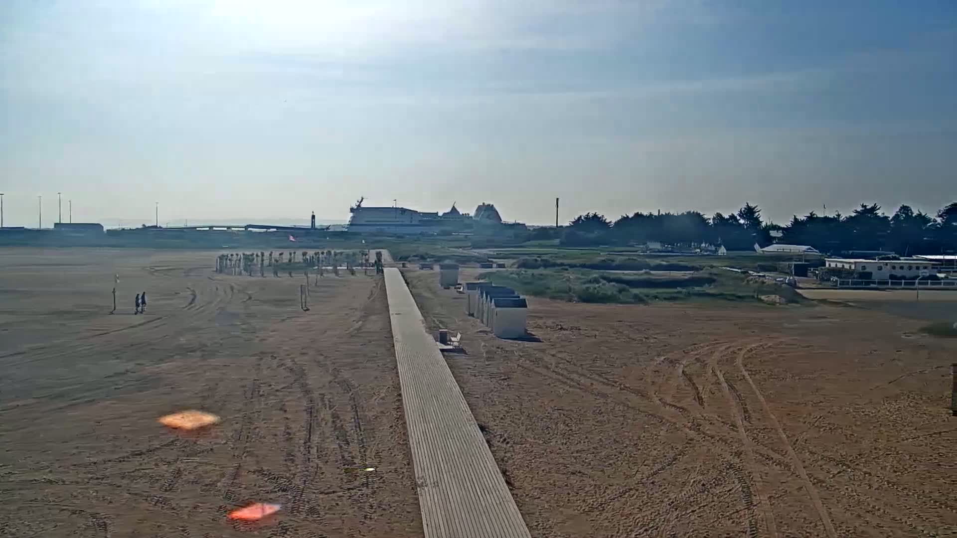 A long wooden boardwalk stretches across a sandy beach towards a distant ferry and a line of trees under a bright, hazy sky.