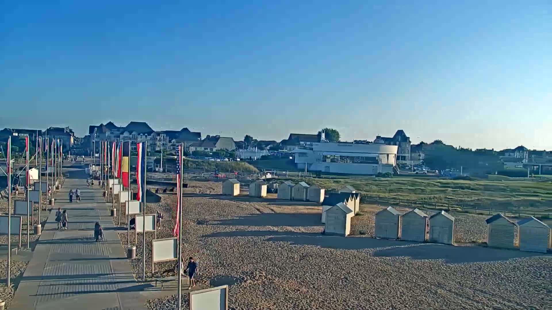 A boardwalk lined with flags leads to a sandy beach with rows of small beach huts under a clear blue sky.
