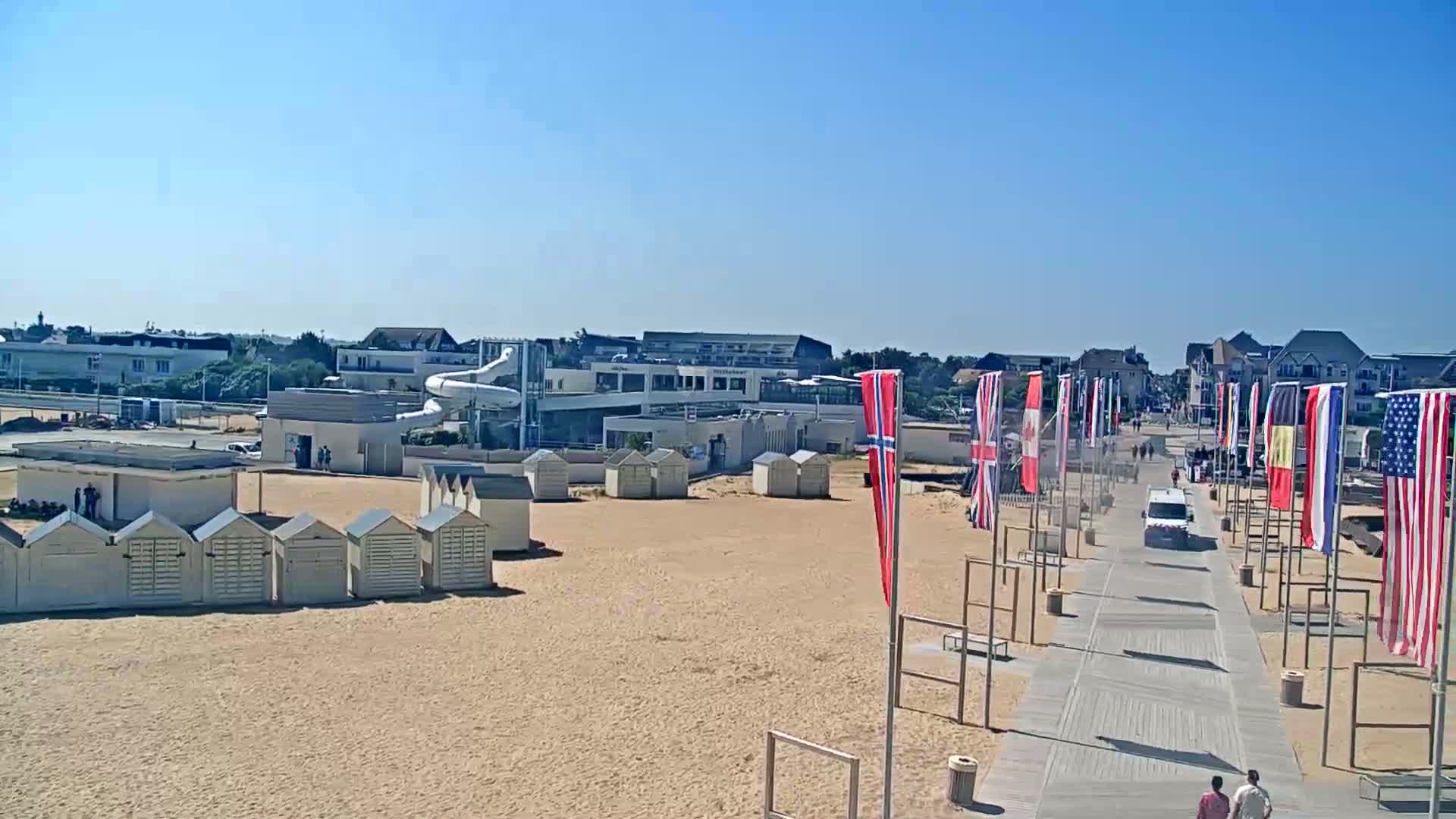 A sandy beach with rows of beach huts, a water slide, and a boardwalk lined with various flags under a clear blue sky.