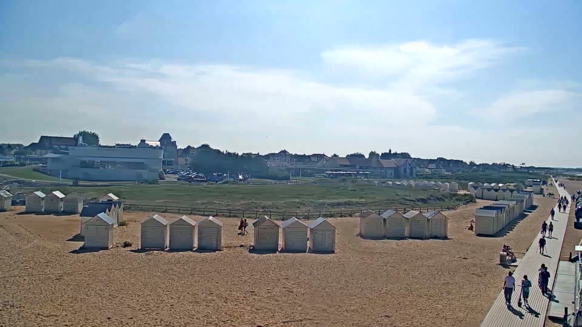 A sandy beach with rows of white beach huts, a boardwalk with people walking on it, and buildings in the background under a mostly sunny sky.