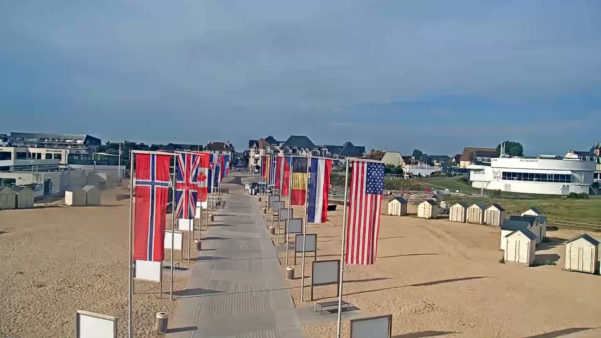 A boardwalk lined with various national flags leads to a sandy beach with small beach huts and buildings in the background under a clear sky.