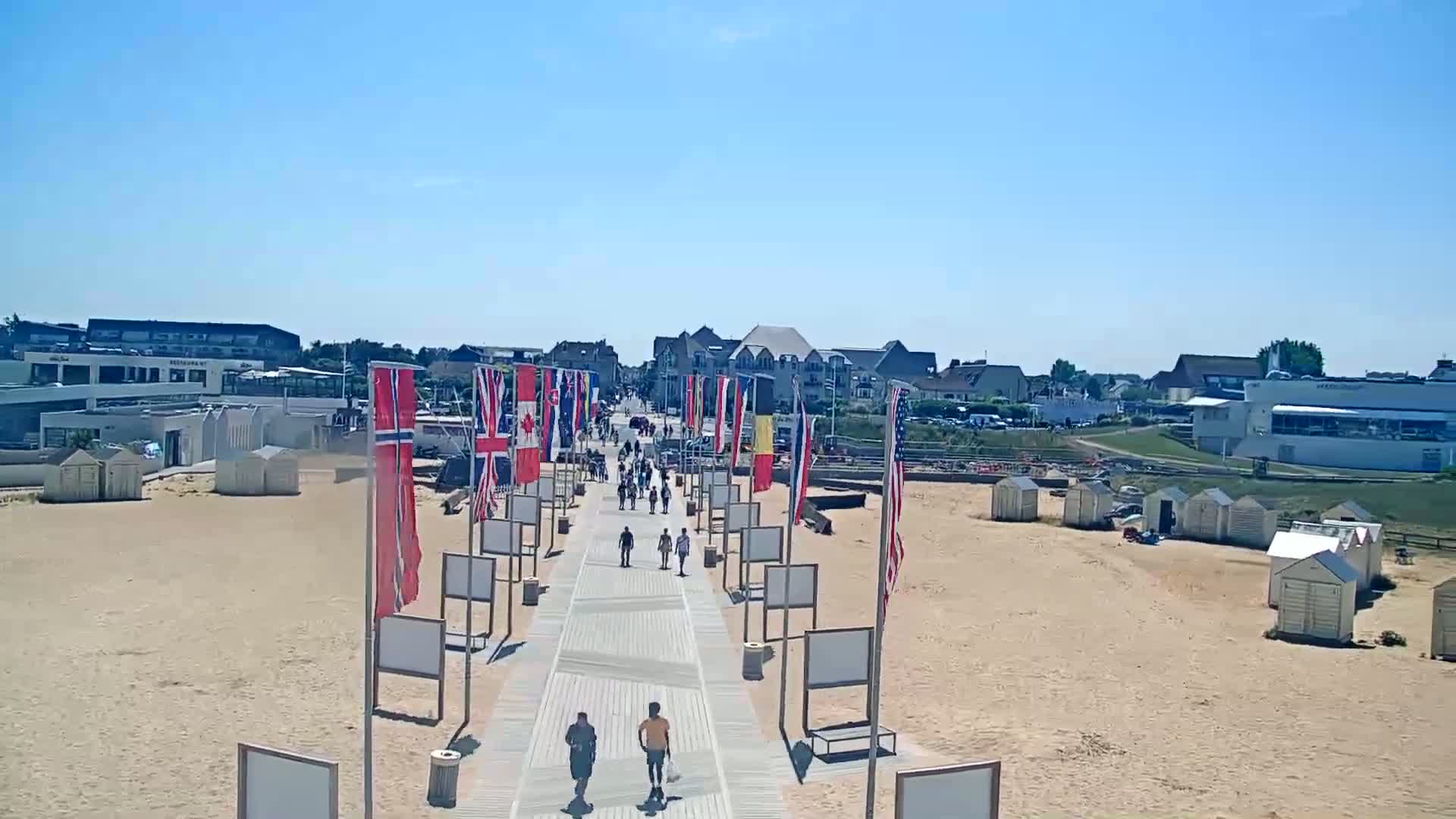 A boardwalk lined with flags leads to a sandy beach with beach huts under a clear blue sky.