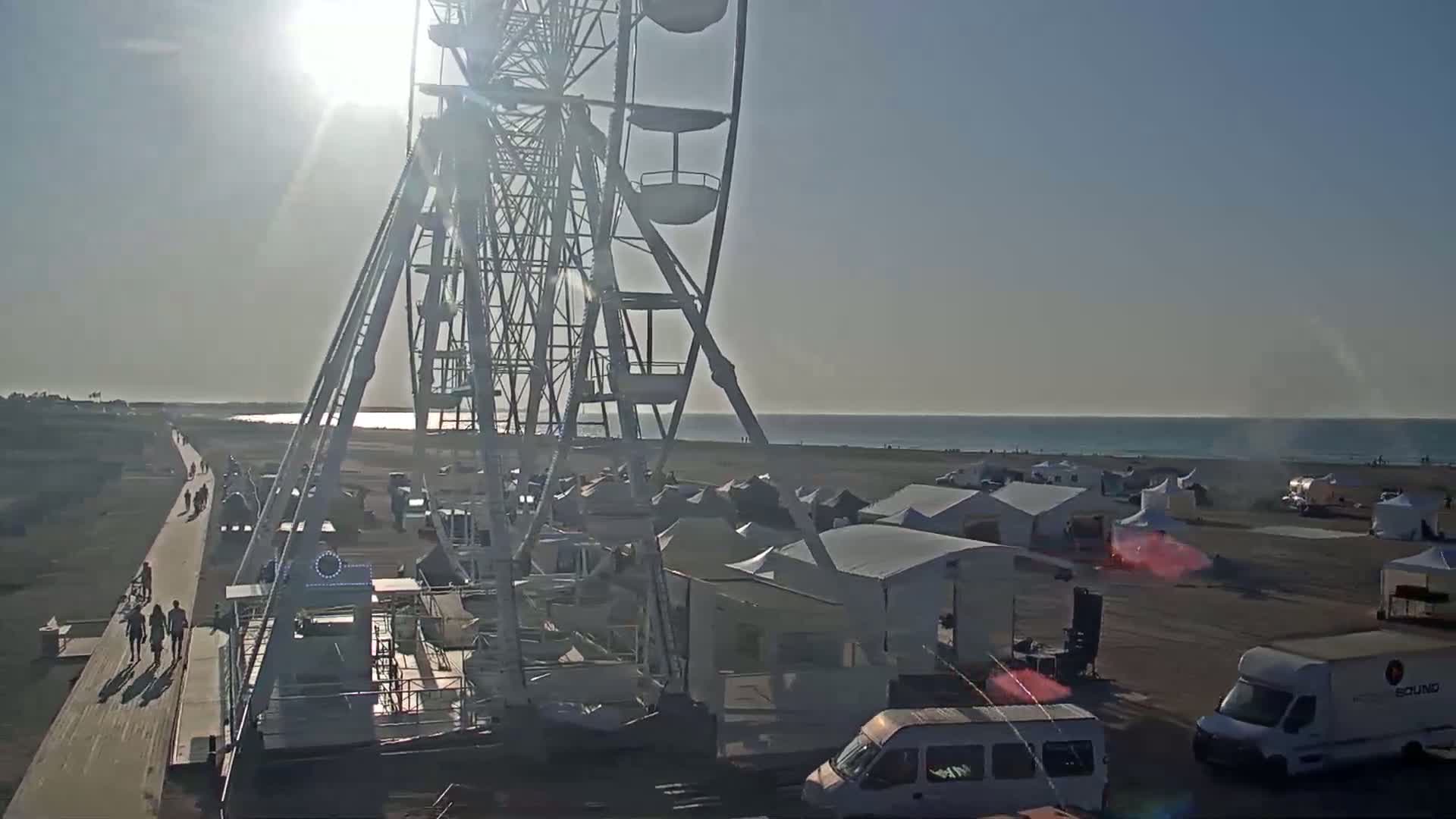 A Ferris wheel is situated near a beach on a sunny day, with numerous tents and vehicles spread across the sand.