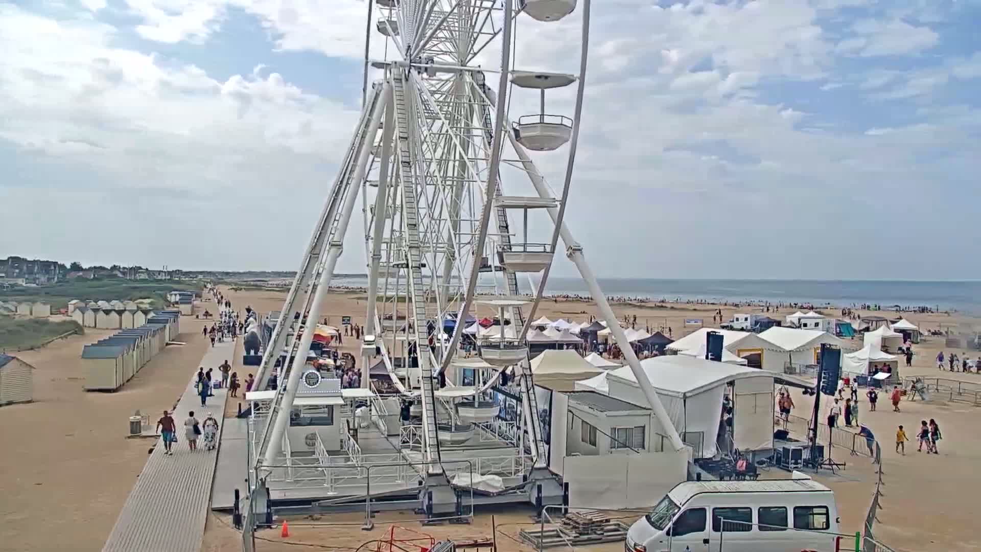 A large Ferris wheel sits on a sandy beach next to a boardwalk and temporary tents under a partly cloudy sky.