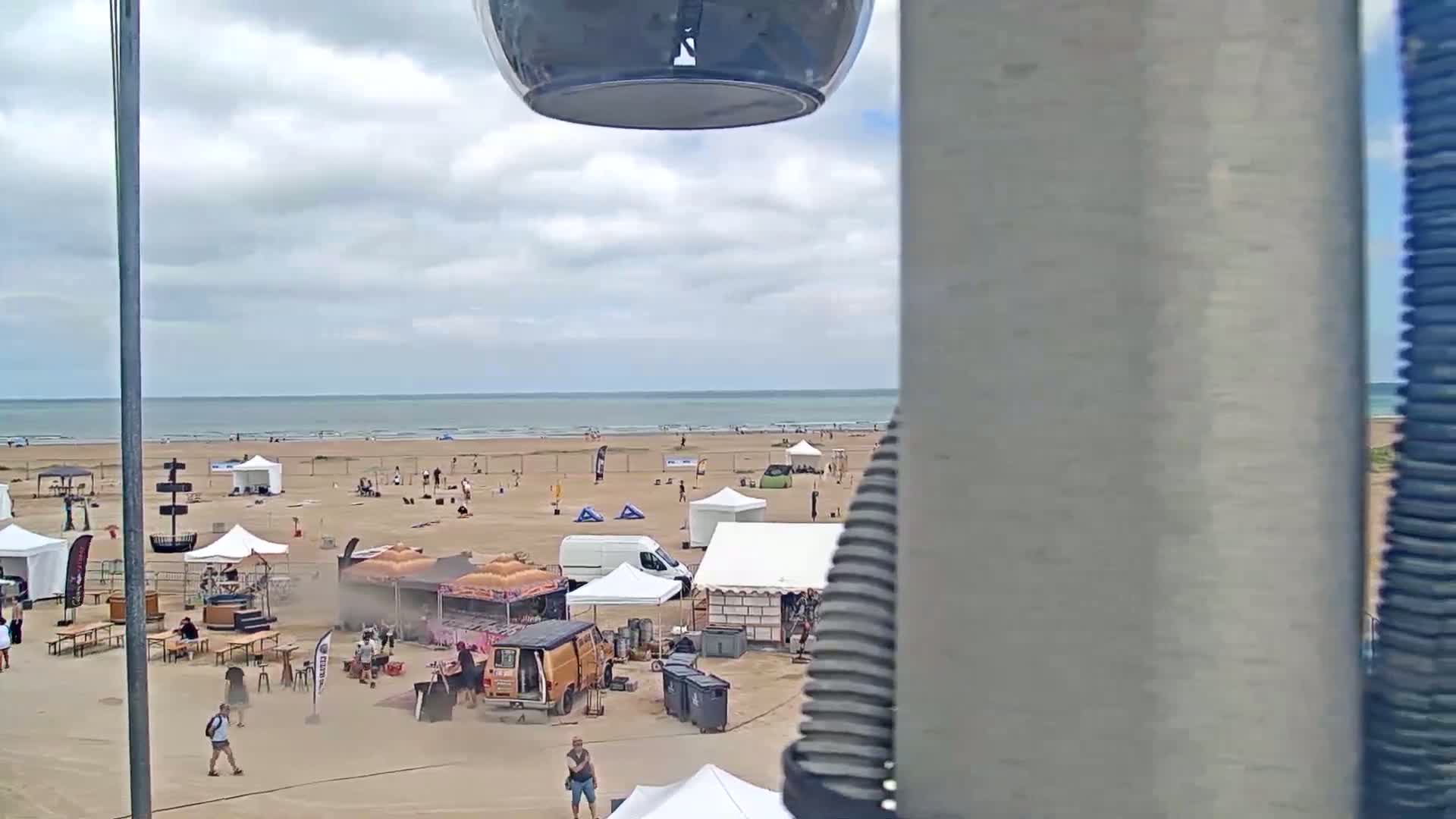 A beach with numerous tents, vendors, and people is visible under a partly cloudy sky, overlooking a calm ocean.