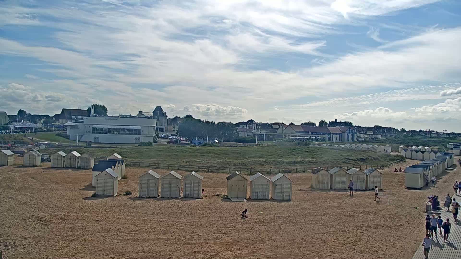 A sandy beach with rows of small beach huts stretches toward a seaside town under a partly cloudy sky.