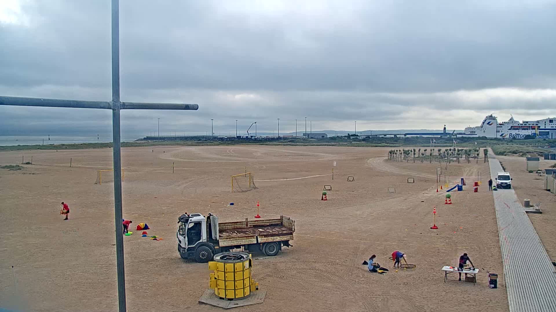 A sandy beach with various recreational activities set up, including soccer goals and cones, a truck, and people, under an overcast sky and near a body of water with a large ferry in the distance.