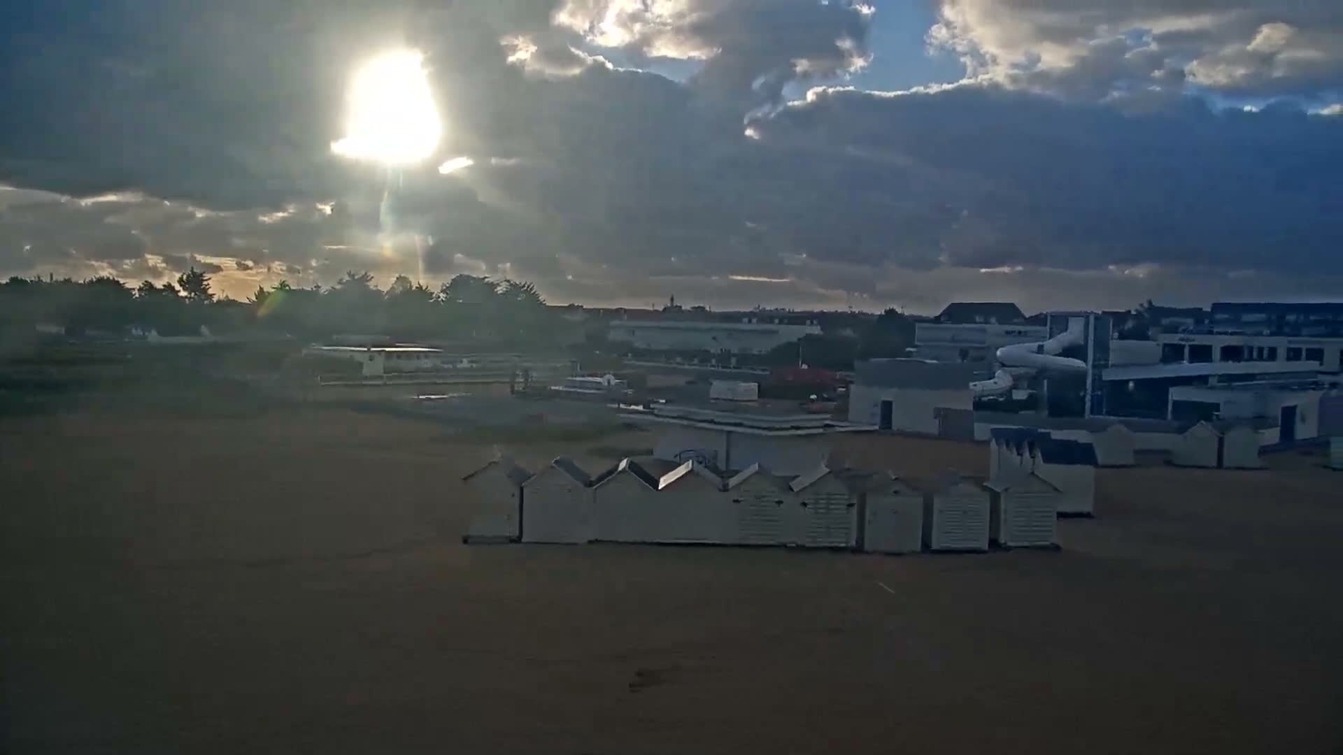 A bright low sun shines through dramatic, partly cloudy skies over a beach with rows of white huts in the foreground and a town with various buildings, including one with a water slide, in the distance.
