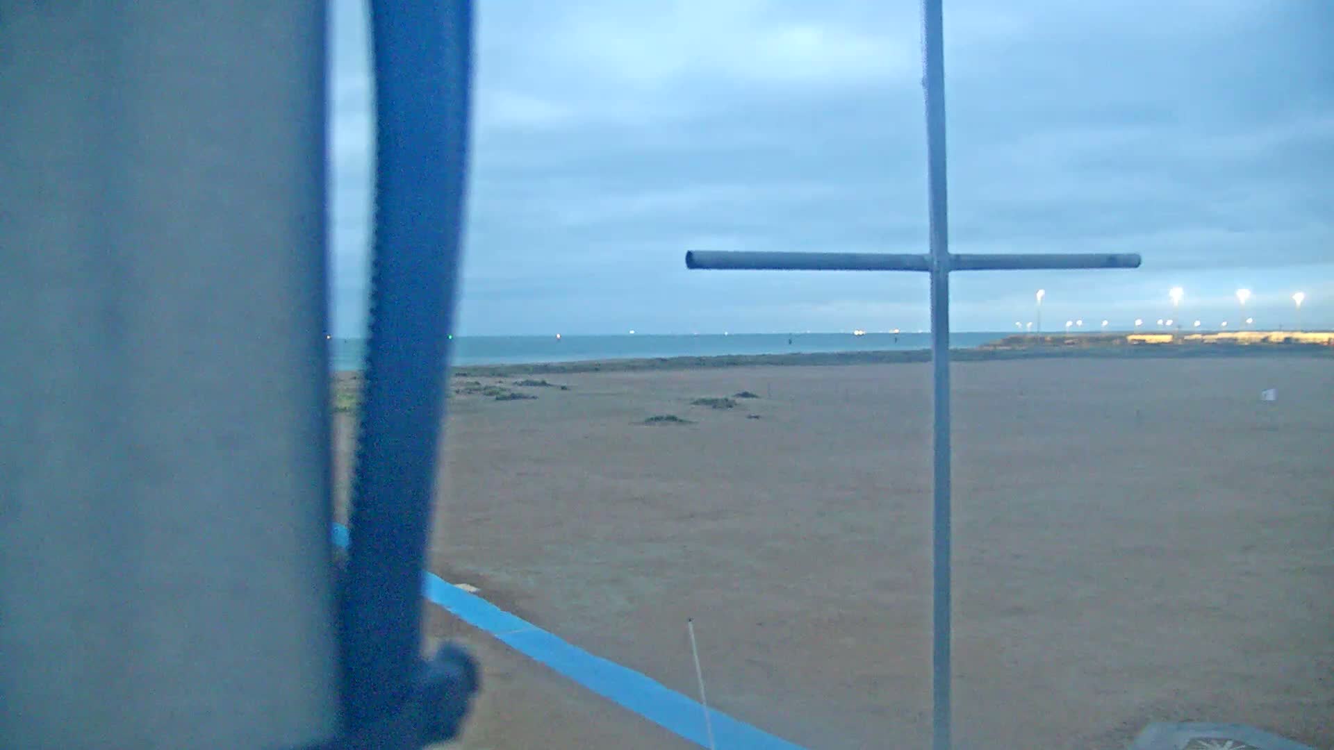 An overcast dusk or dawn scene reveals a sandy beach meeting a calm sea with distant port lights under a cloudy sky, partially framed by a blue foreground structure and a grey cross-shaped pole.