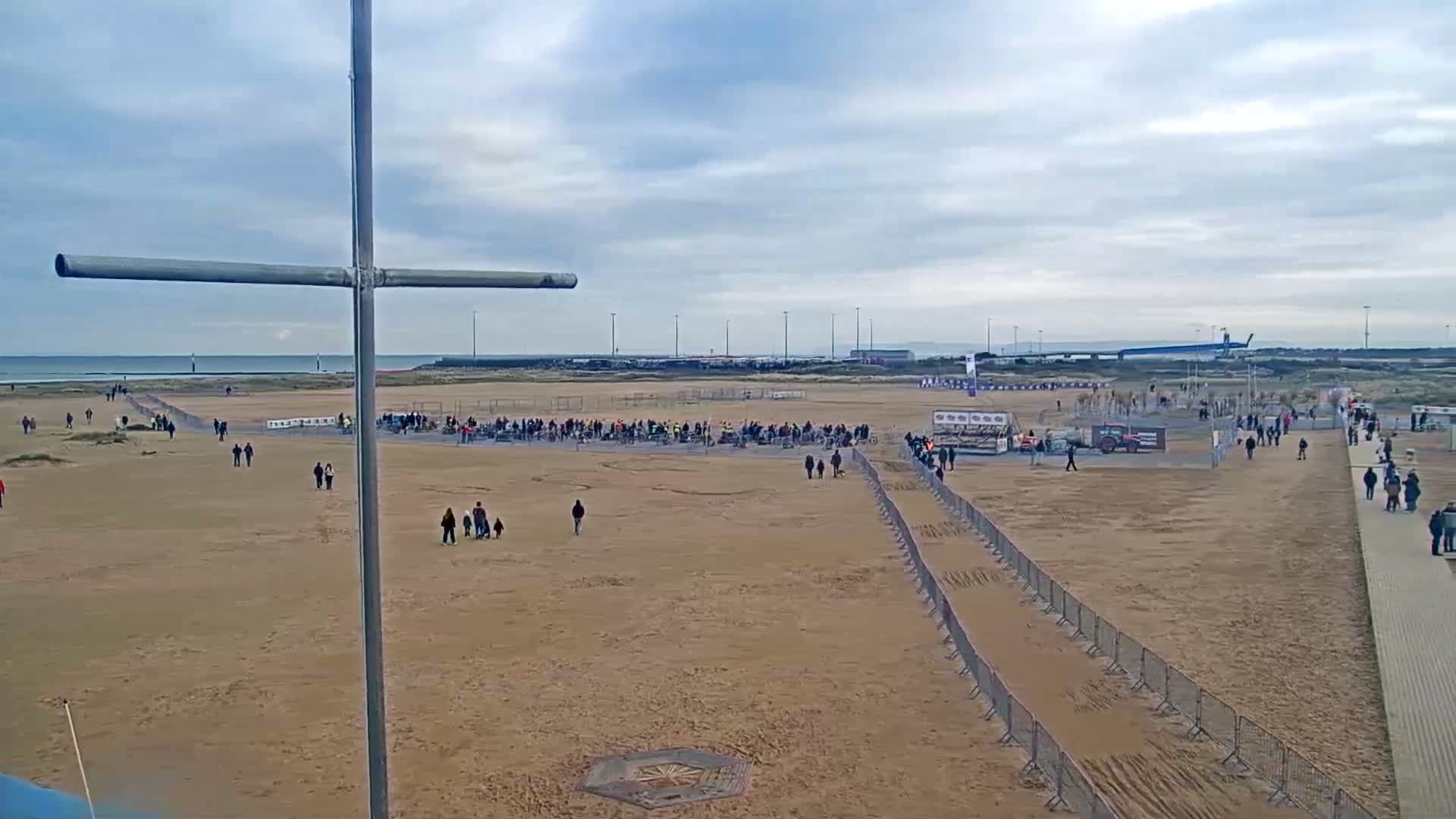 An overcast dusk or dawn scene reveals a sandy beach meeting a calm sea with distant port lights under a cloudy sky, partially framed by a blue foreground structure and a grey cross-shaped pole.